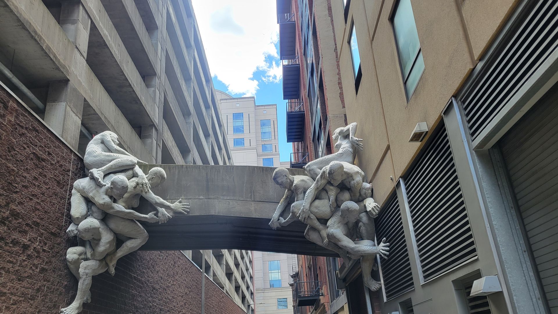 A sculpture of bodies holding up a pedestrian foot bridge outside an alley across from Reading Terminal Market. 