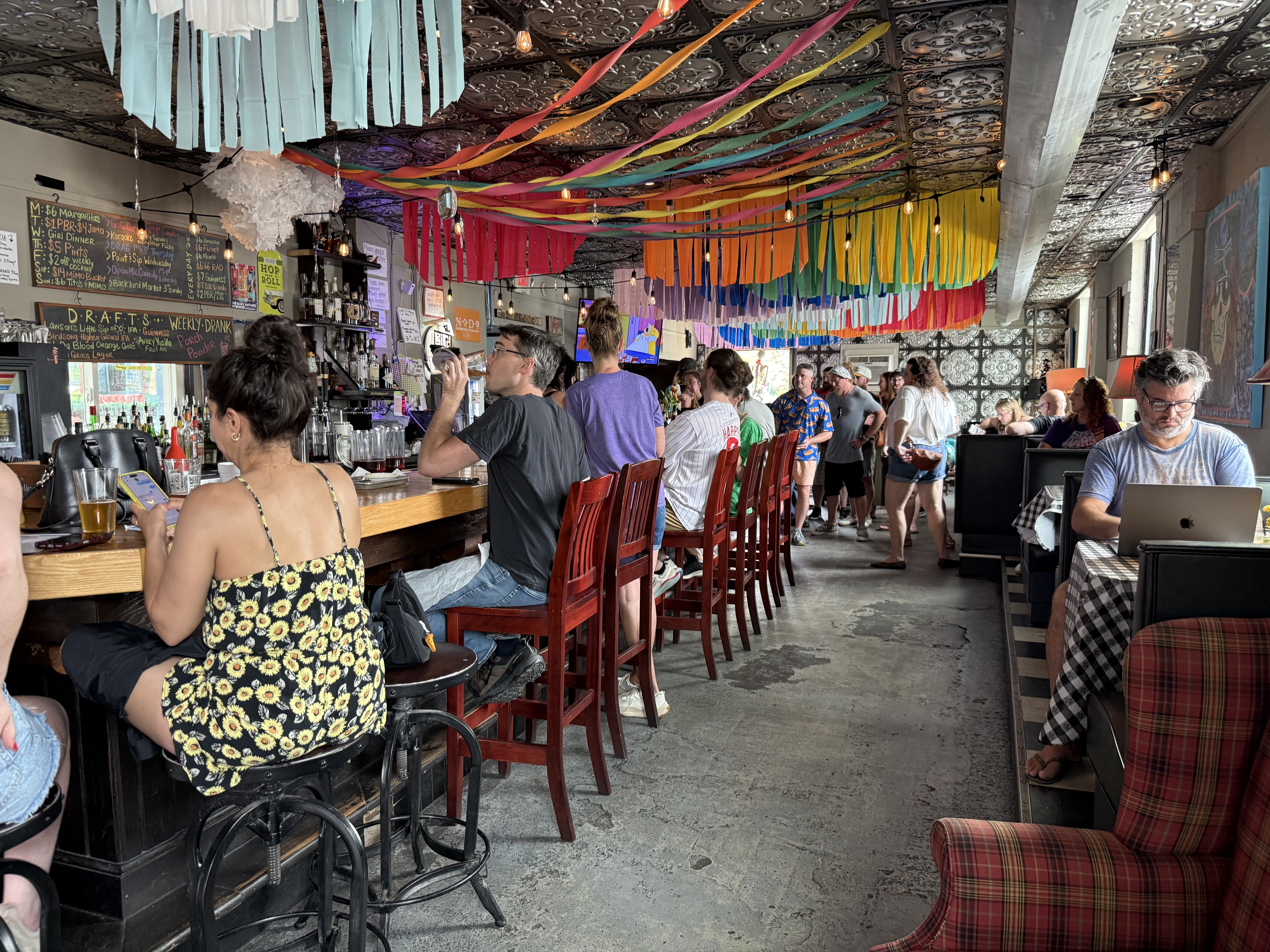 A busy bar interior with a long left-side counter and red stools, rainbow streamers across a patterned ceiling, and groups of people chatting, drinking, or working on laptops.
