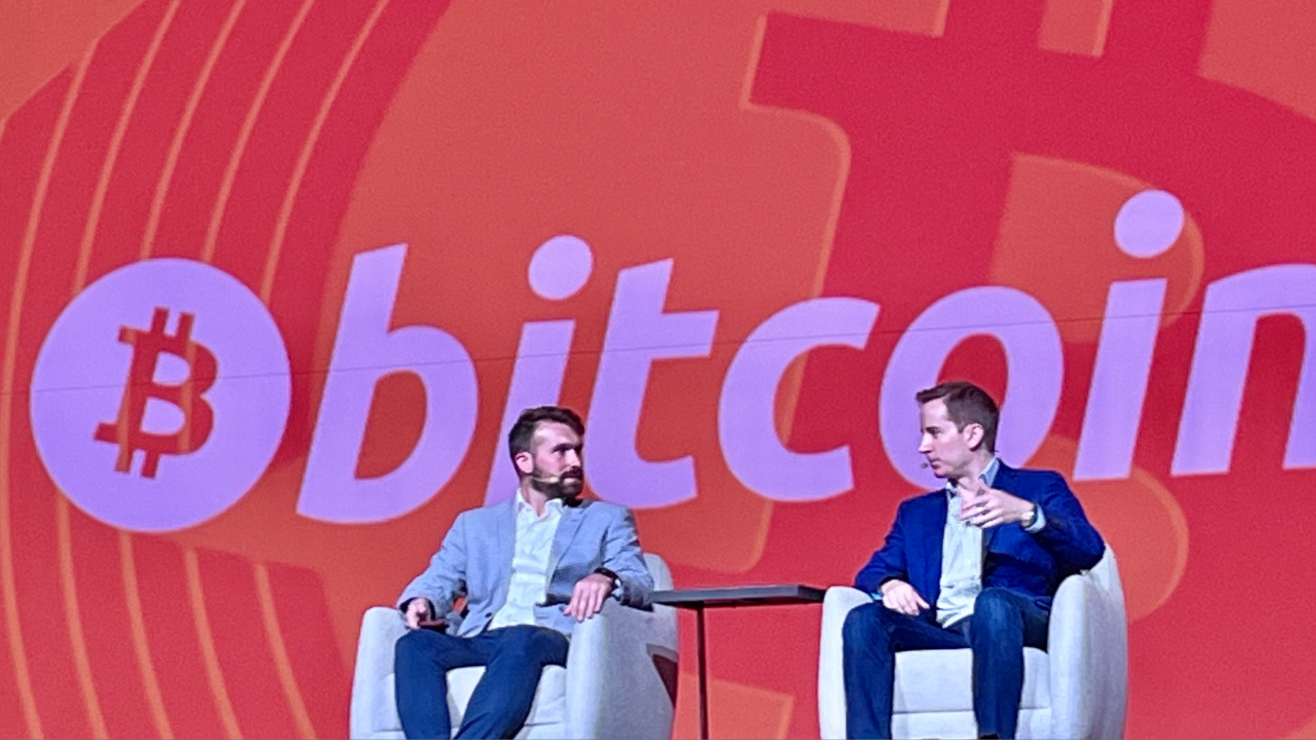 Two men talk in chairs on stage, with a massive "Bitcoin" sign behind them.