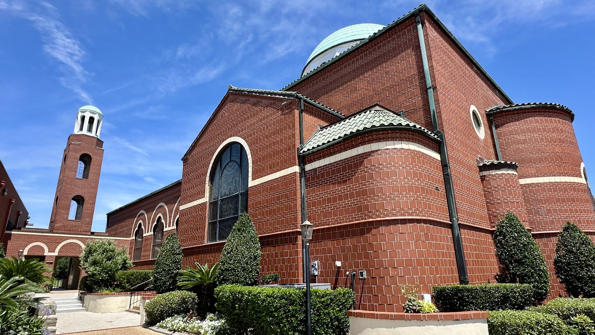 Red brick church with a turquoise dome and tall bell tower, a large arched window, and rounded brick detailing. A neatly landscaped foreground with green shrubs and a clear blue sky.