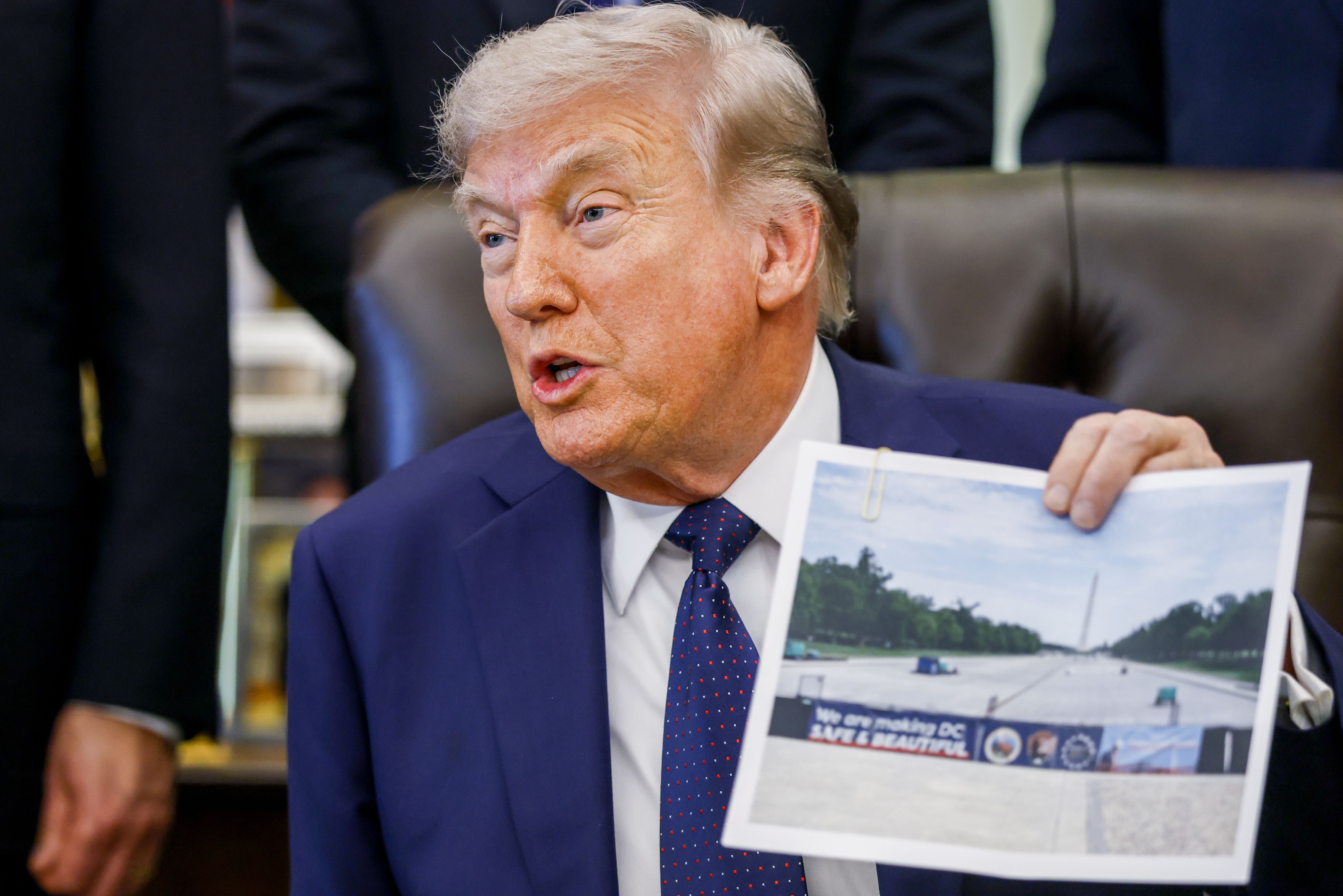 President Donald Trump holds a photo of the Lincoln Memorial reflecting pool with a fence reading "We are making DC Safe & Beautiful" during a healthcare affordability event in the Oval Office of the White House.