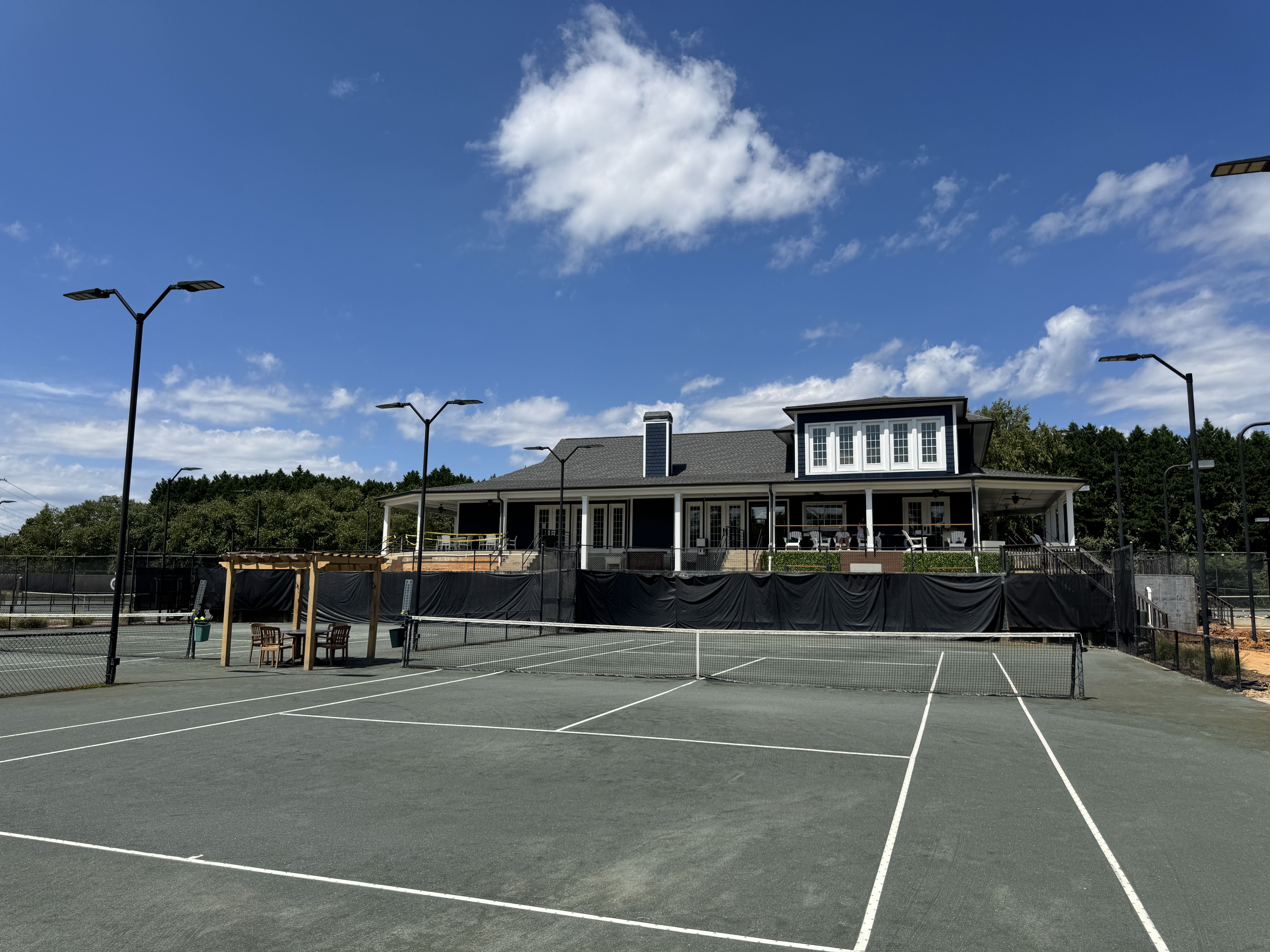 Empty outdoor tennis court with white lines and net, surrounded by black fencing, wooden chairs under a small wooden pergola, and a large clubhouse behind under a blue sky with scattered clouds.
