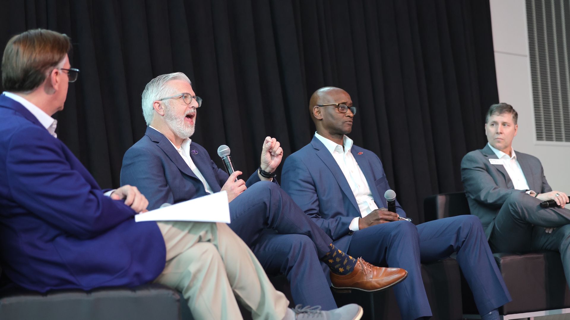 Four men sit on a stage in a panel, each holding a microphone.