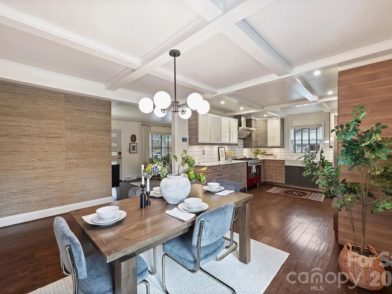 Modern kitchen and dining area with wooden floors, a wooden table set with white dishes, blue velvet chairs, white coffered ceiling, and a mix of wood and white cabinets.