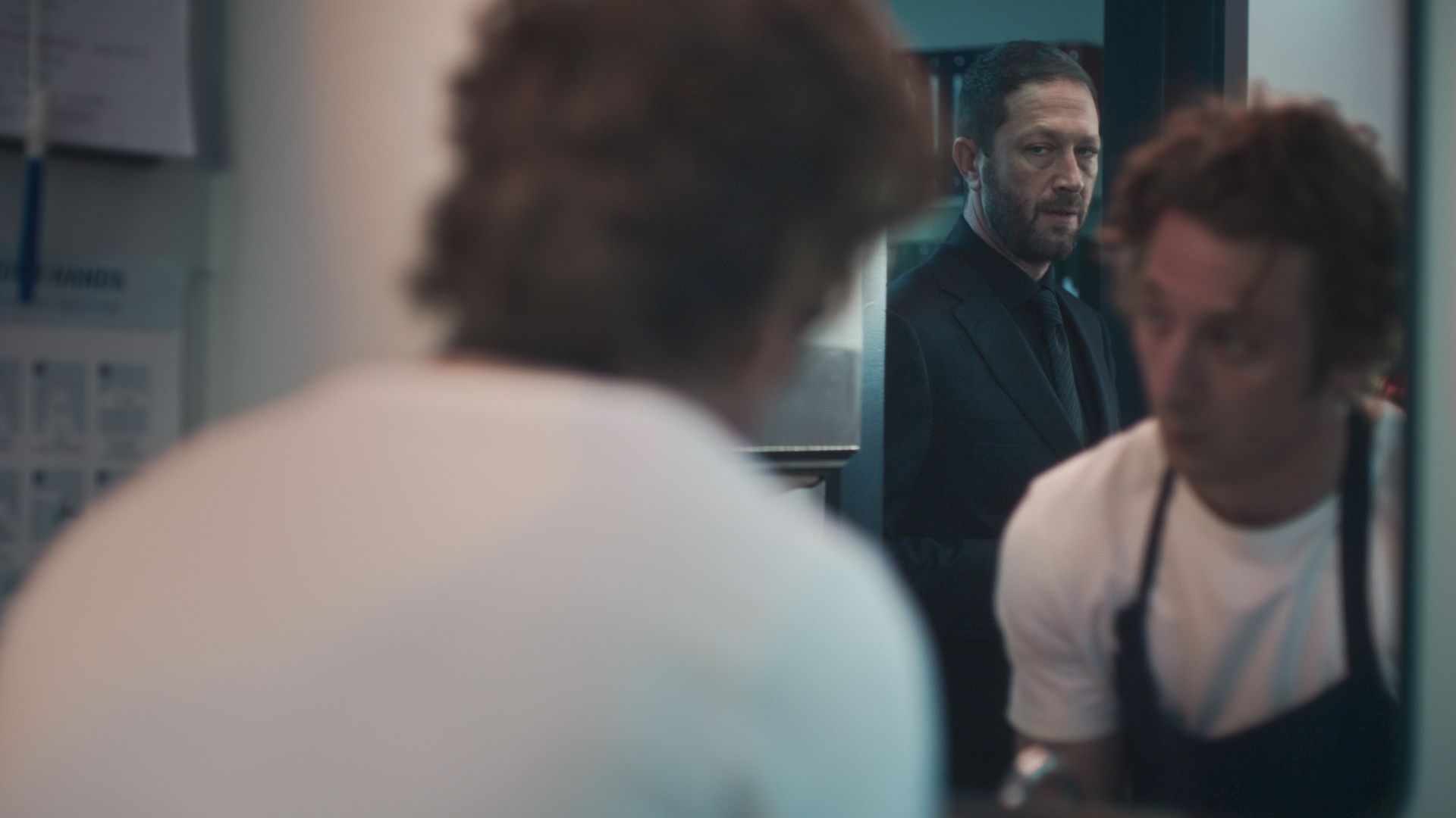 Man in black suit and tie looks intently while standing in a dimly lit room; another man with curly hair in white shirt and black apron is blurred in the foreground.