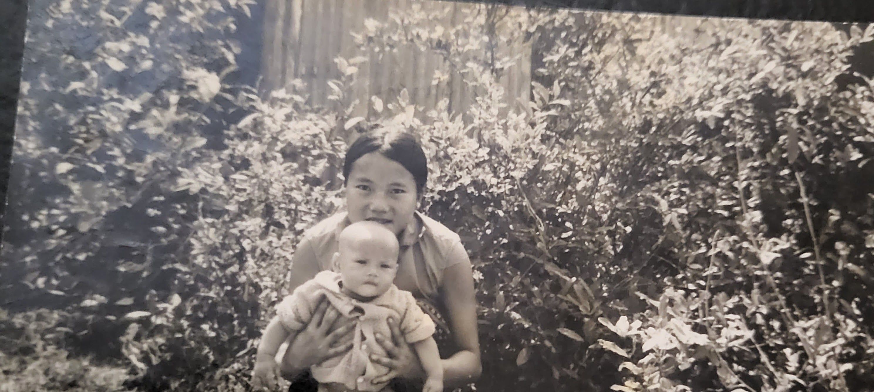 An old black and white photo shows a baby and her mother in front of some greenery.