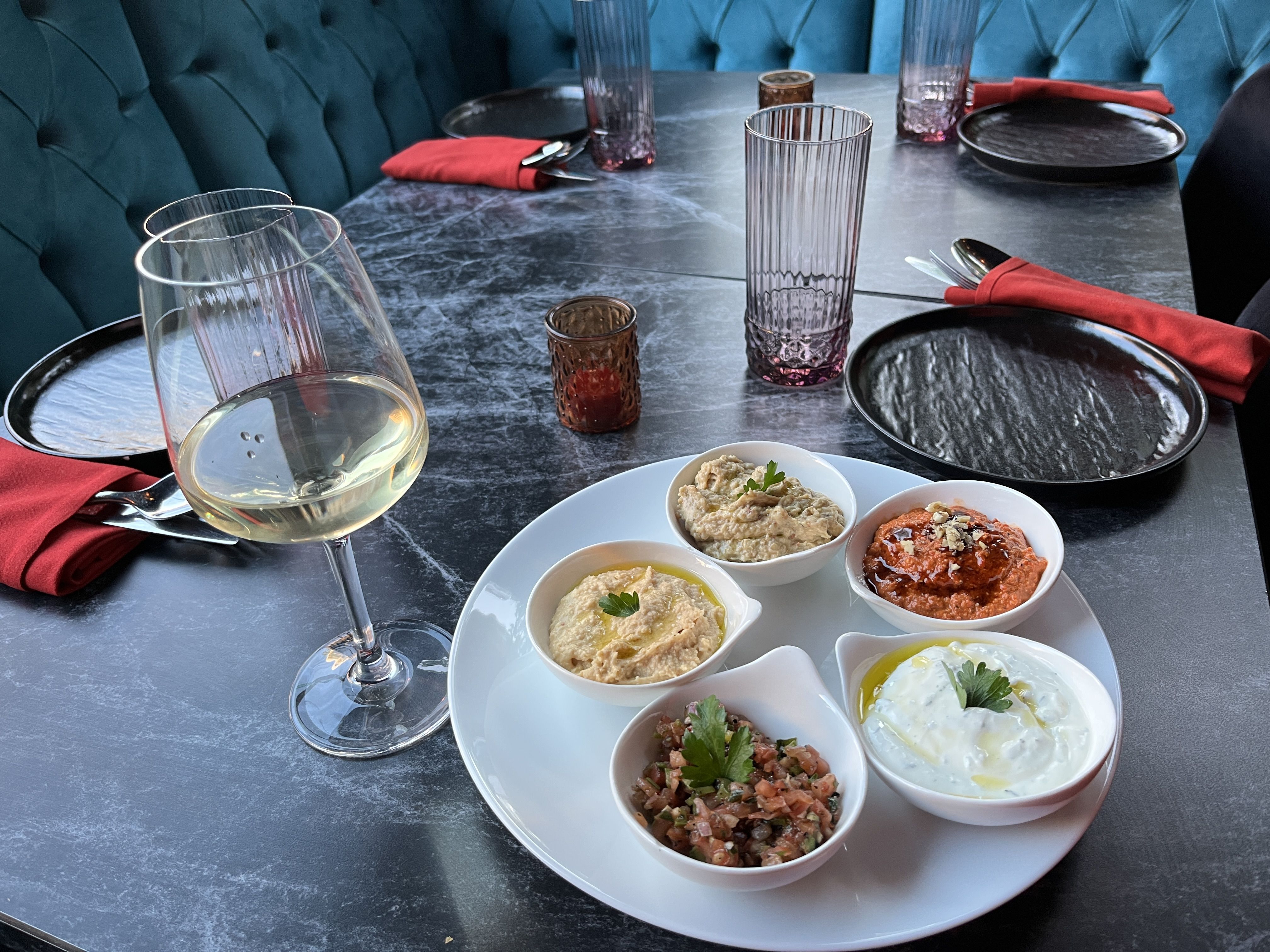 Table set with black plates, red napkins, and purple glasses; a white wine glass and a white plate hold five bowls of colorful dips garnished with herbs on a dark marble surface.
