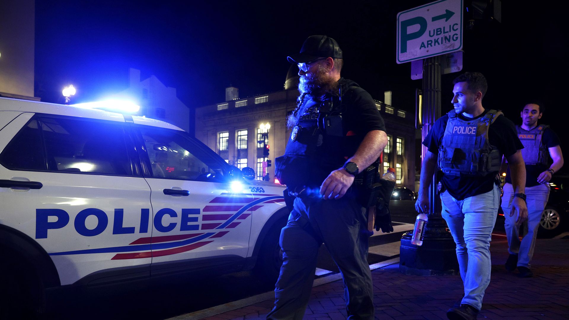 Three police officers in tactical gear walking near a marked police SUV with flashing blue lights at night on a city sidewalk under a public parking sign.