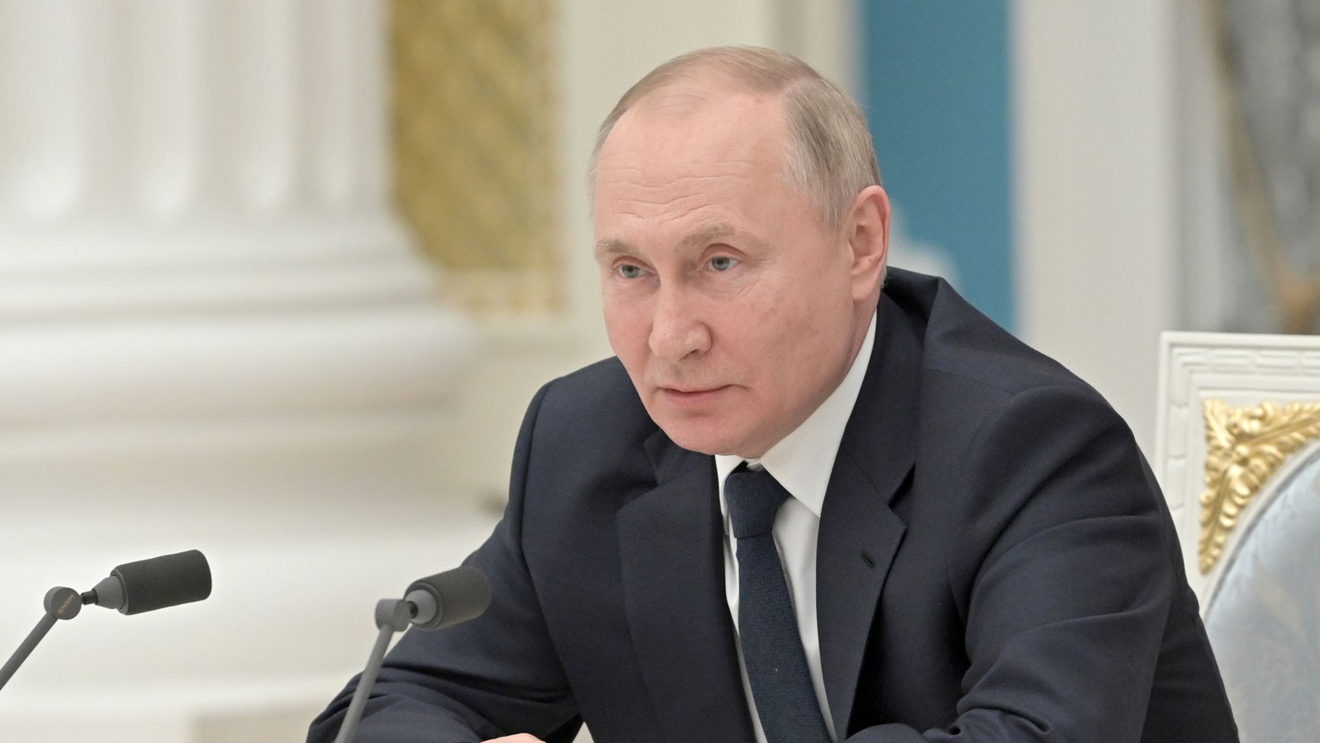 Photo of Vladimir Putin sitting with his hands folded in front of him as he leans forward toward a mic