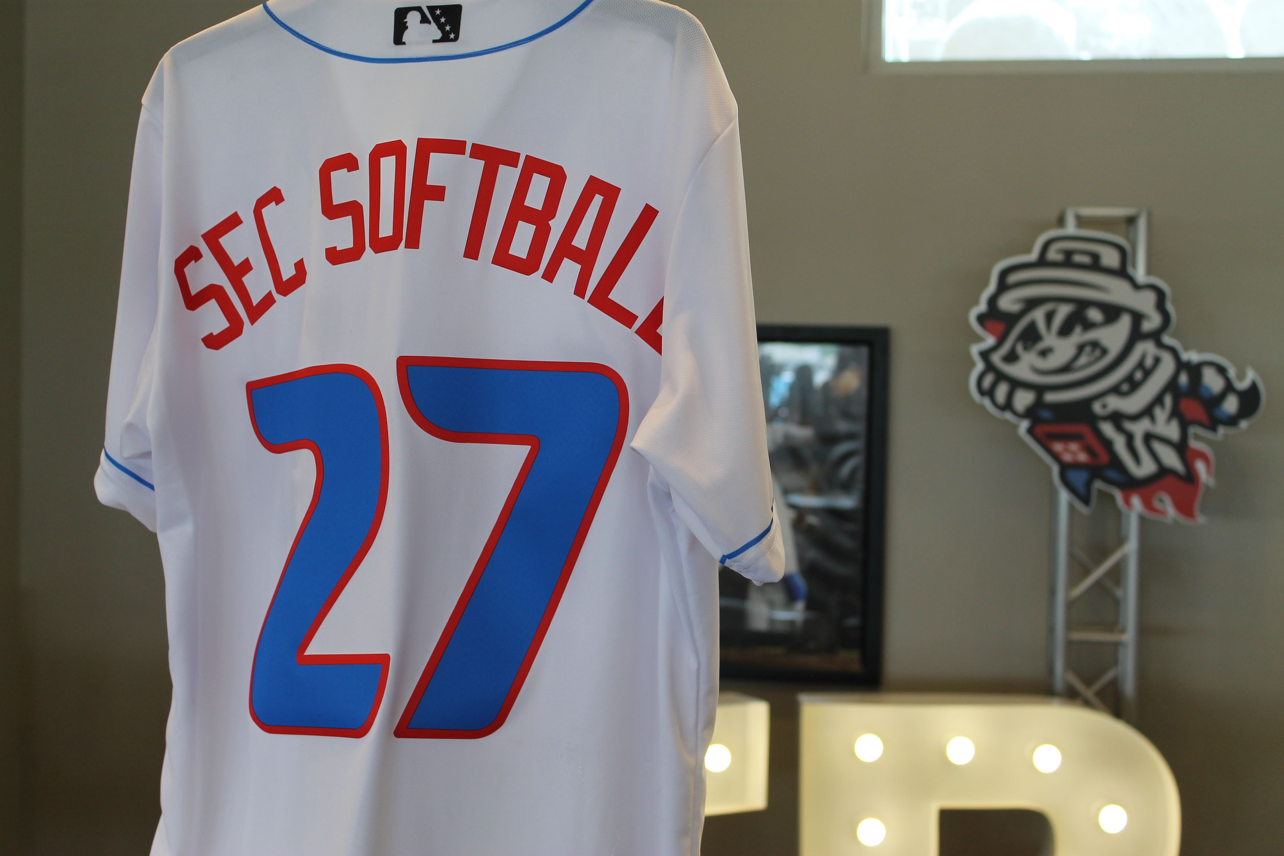 Back view of a white softball jersey with red text reading "SEC SOFTBALL" and a large blue number 27 outlined in red; blue piping on the collar and sleeves; a mascot cutout and lit signage visible in the background.