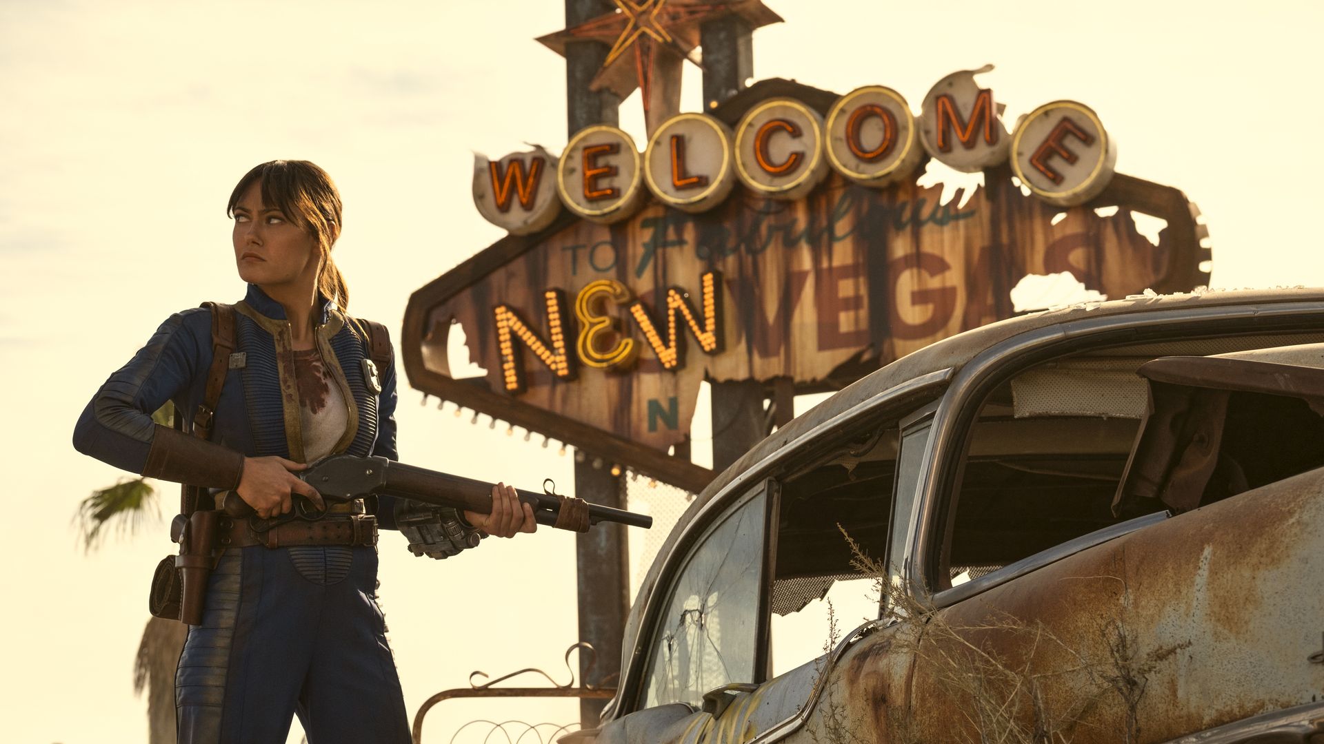 Woman in blue jumpsuit holding shotgun stands beside rusty car with broken windows under faded "Welcome to New Vegas" sign in a desert setting at sunset.