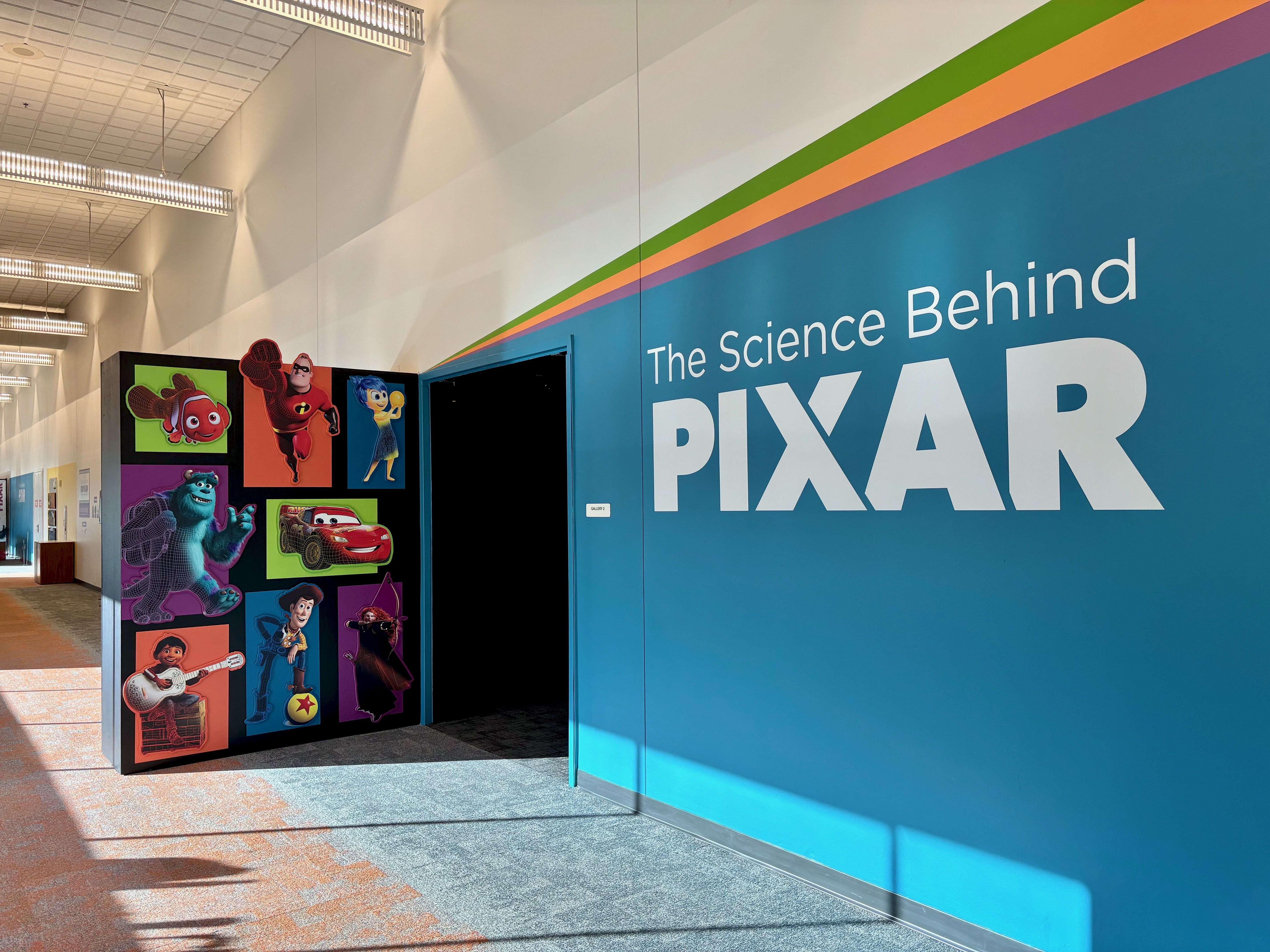 Wall exhibit titled "The Science Behind PIXAR" with colorful stripes above text and images of Pixar characters like Nemo, Mr. Incredible, Joy, Sulley, Lightning McQueen, Miguel, Woody, and Merida on a black panel.