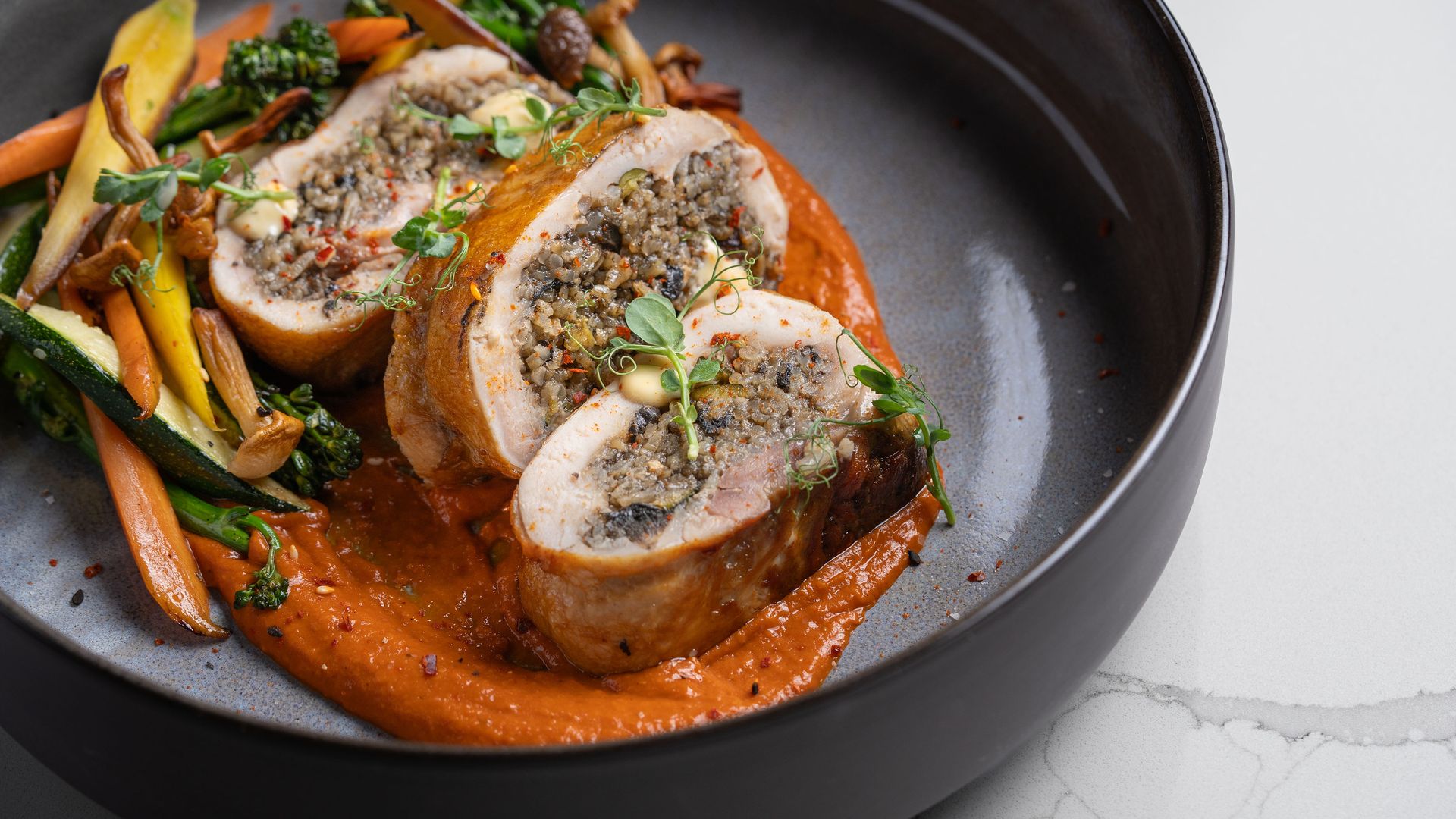 Three slices of stuffed chicken on red sauce, garnished with microgreens, served with assorted sautéed vegetables in a dark bowl on a white marble surface.