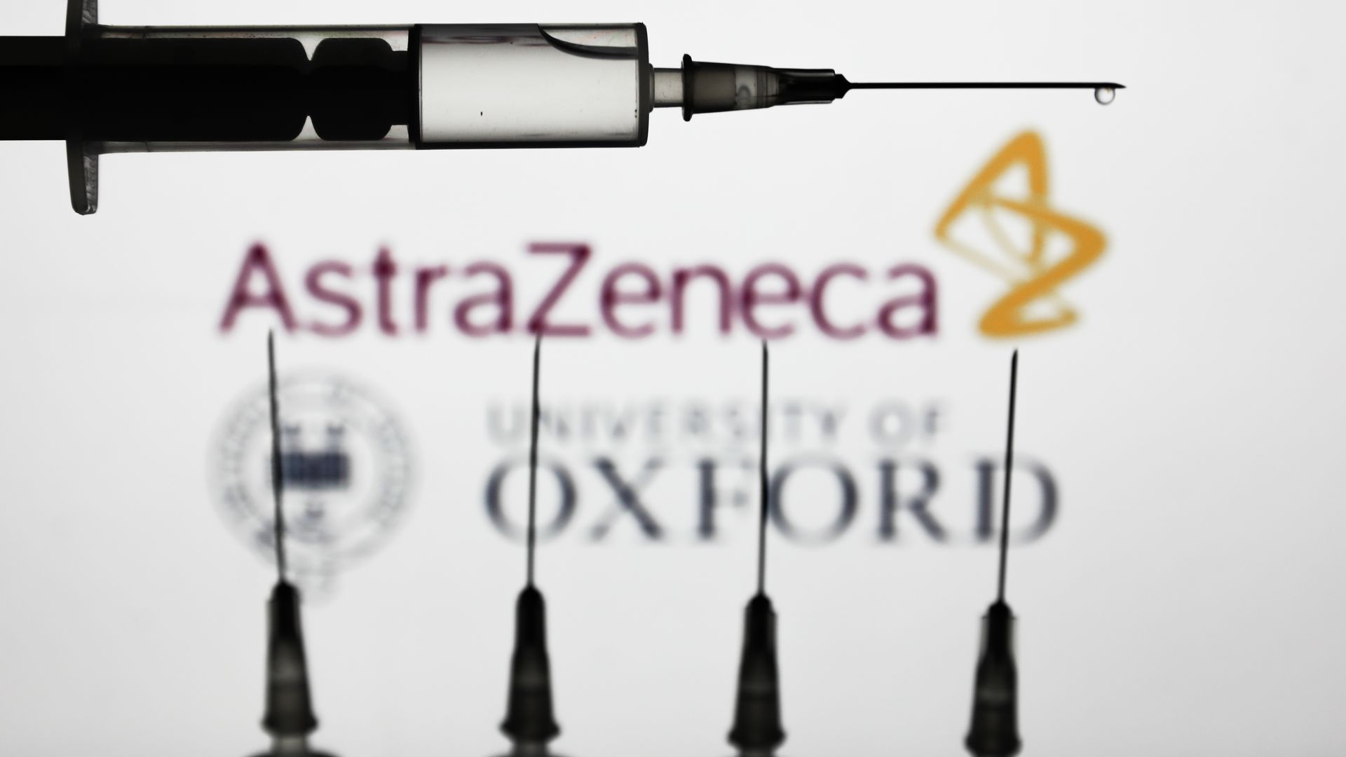 Medical syringes are seen with AstraZeneca and University of Oxford logos displayed on a screen in the background in this illustration photo taken in Poland