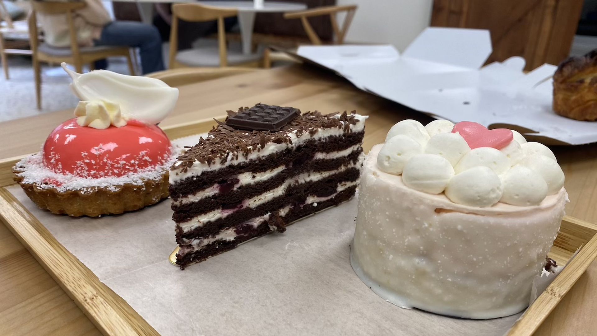 Three assorted desserts on a wooden tray: a red-glazed tart with white cream and coconut, a slice of layered chocolate cake with cream and chocolate shavings, and a small white cake topped with cream and a pink heart.