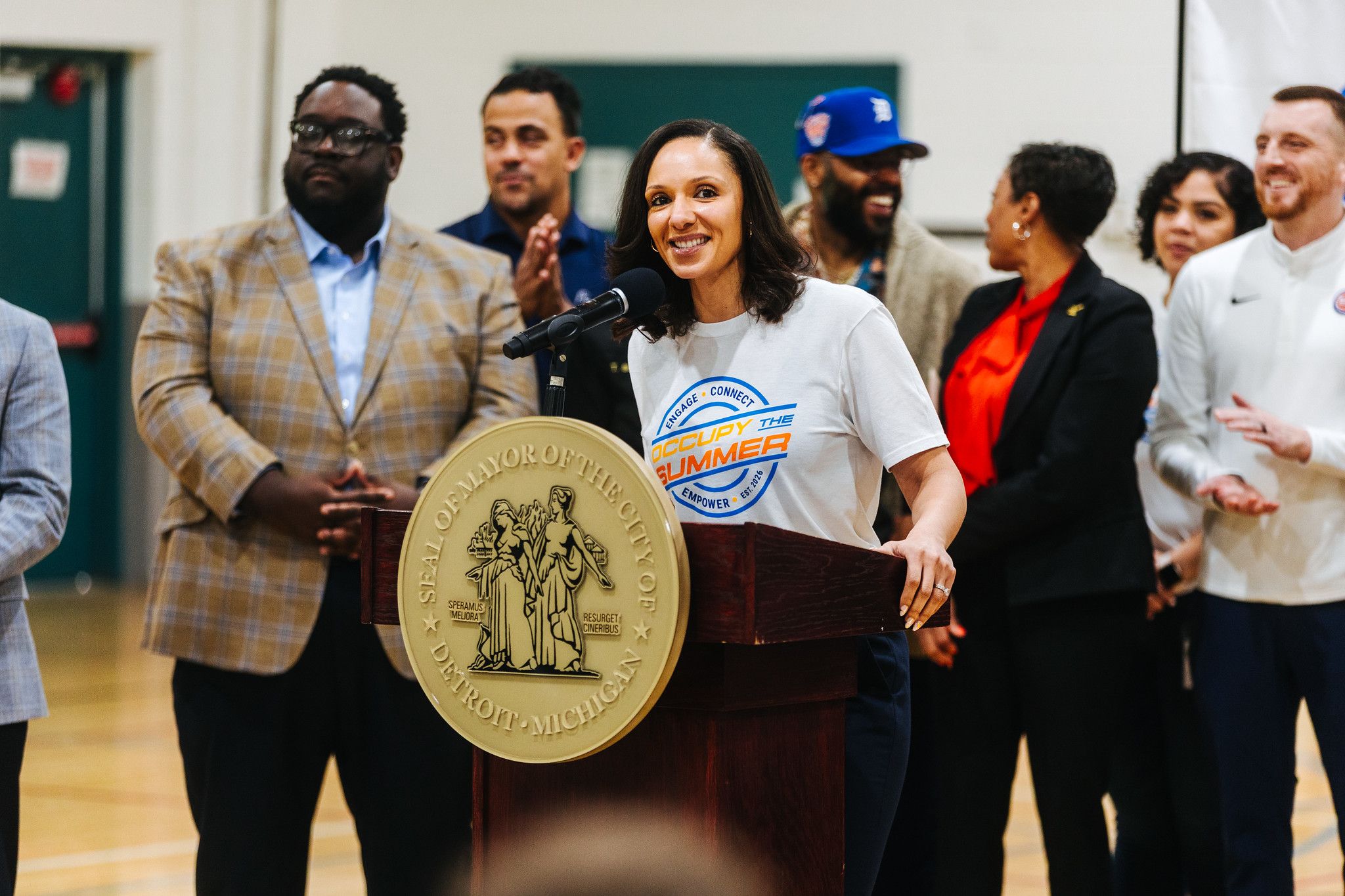 Sheffield in a white T-shirt speaks at a podium with a large gold seal and microphone, as a group in business-casual attire applauds behind her in a gymnasium.