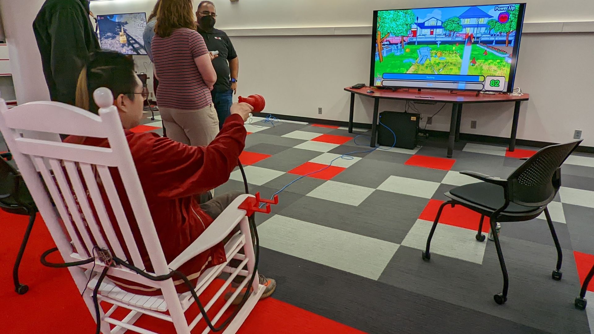 A man plays a video game in a white rocking chair, with a game controller shaped like a hose nozzle. 
