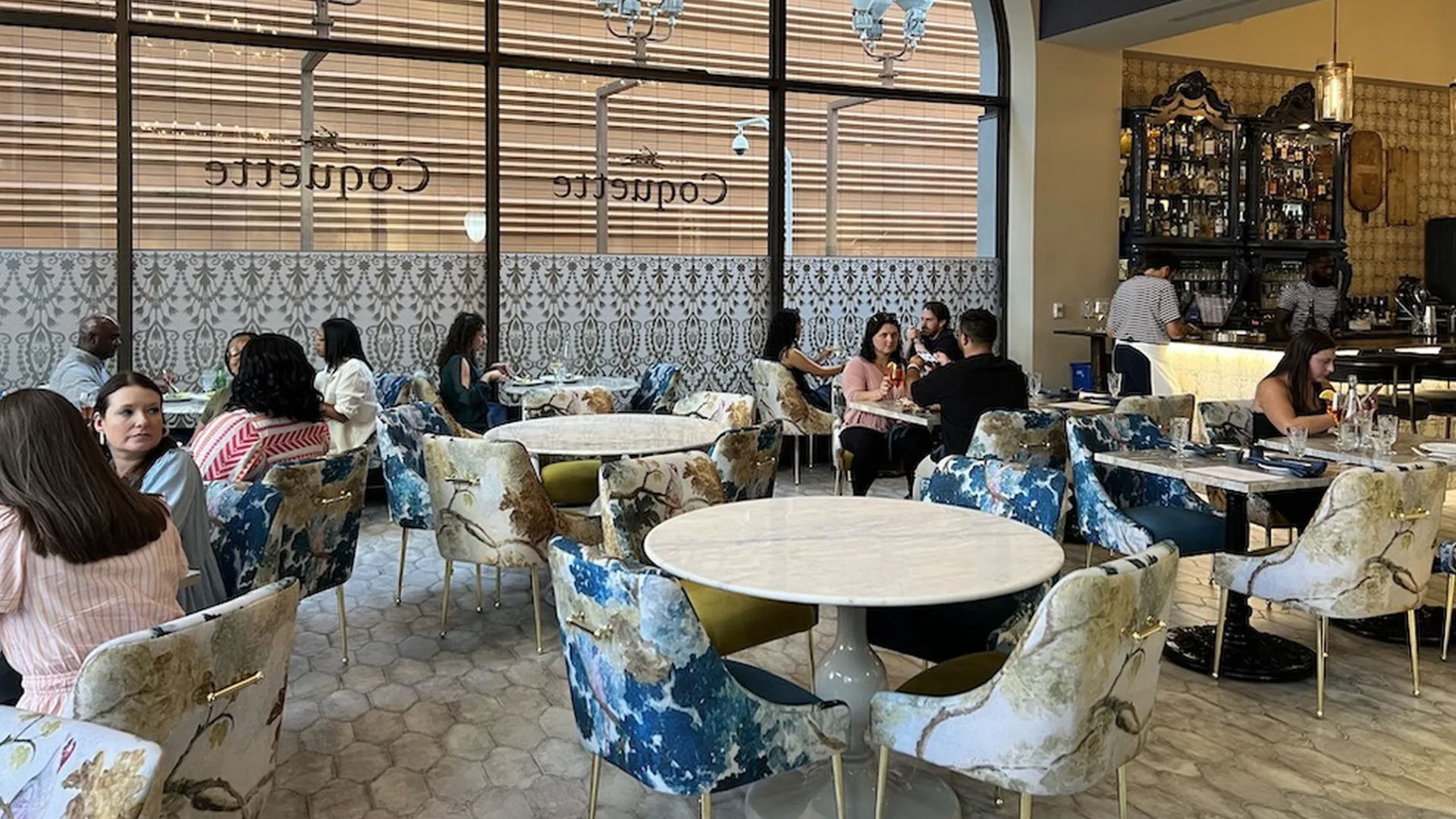 A modern restaurant interior with round white marble tables and floral-patterned chairs in blue, green, and cream colors. Several people are seated, dining and talking. A bar with shelves of bottles is in the background.