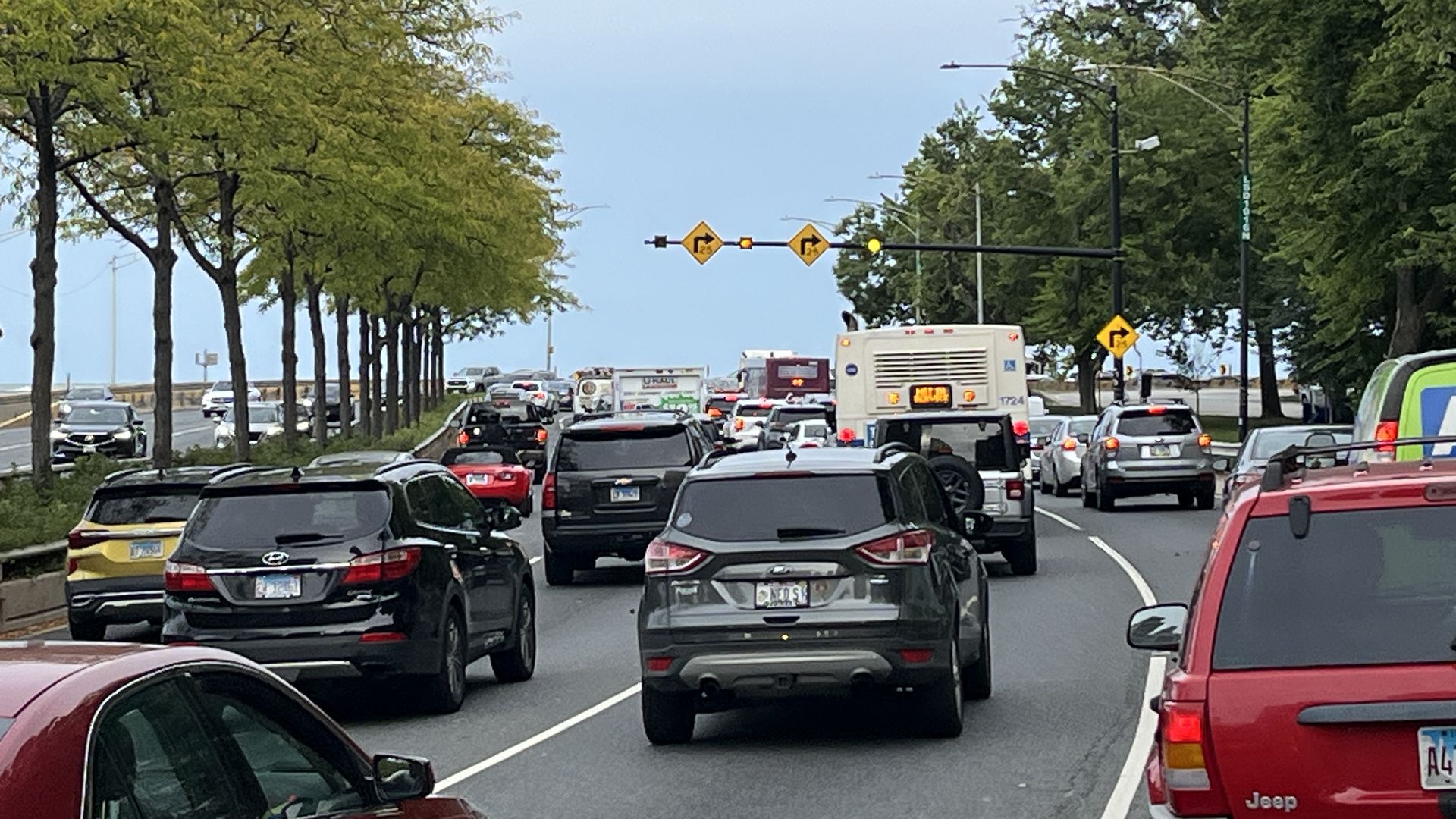 Traffic jam on a tree-lined road with multiple cars and a bus, under cloudy sky and yellow traffic signs indicating right turns ahead.
