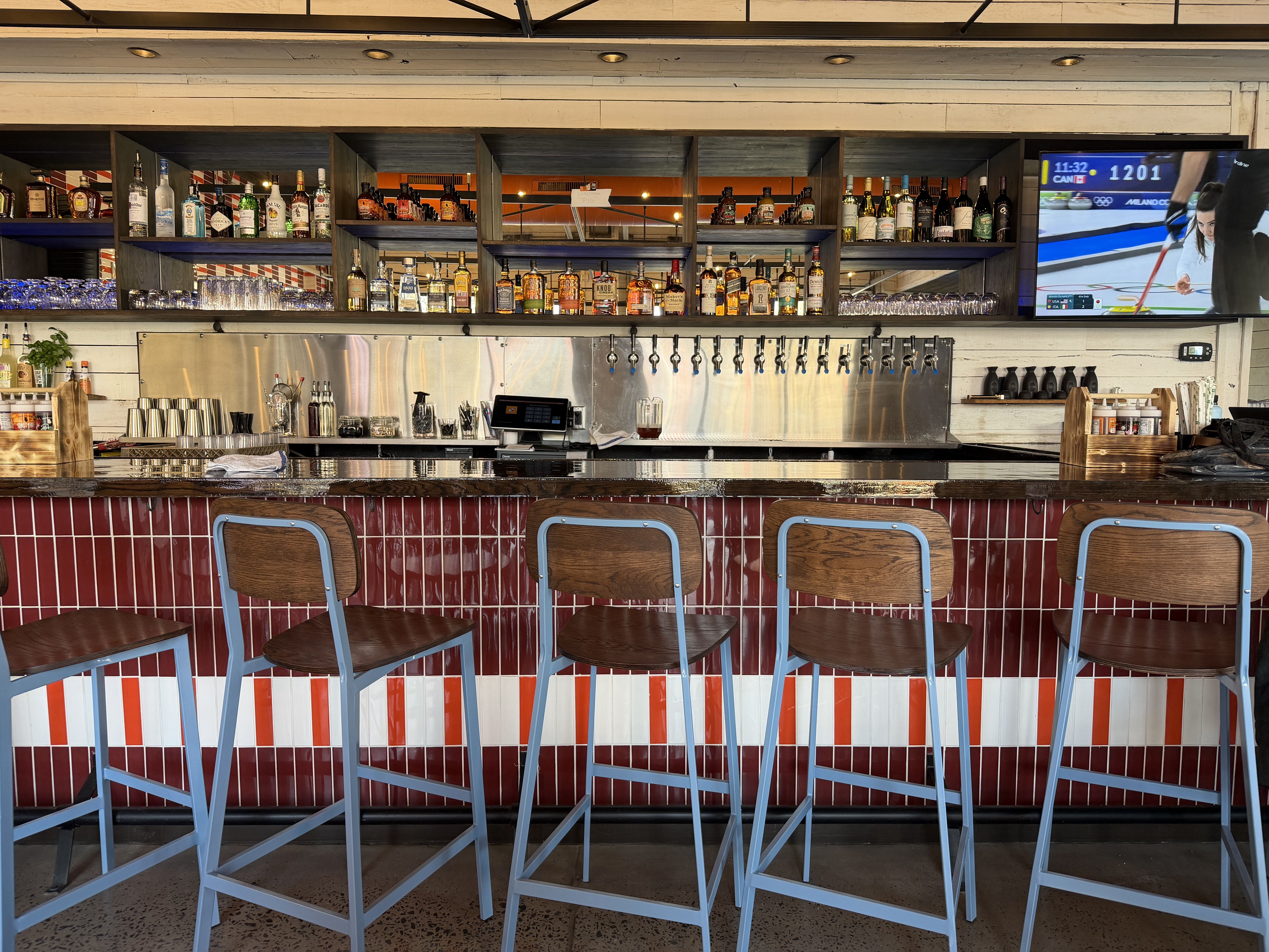 Empty bar with five brown wooden stools with blue metal frames, red and white tiled counter, shelves of liquor bottles, beer taps, and a TV showing a curling match.