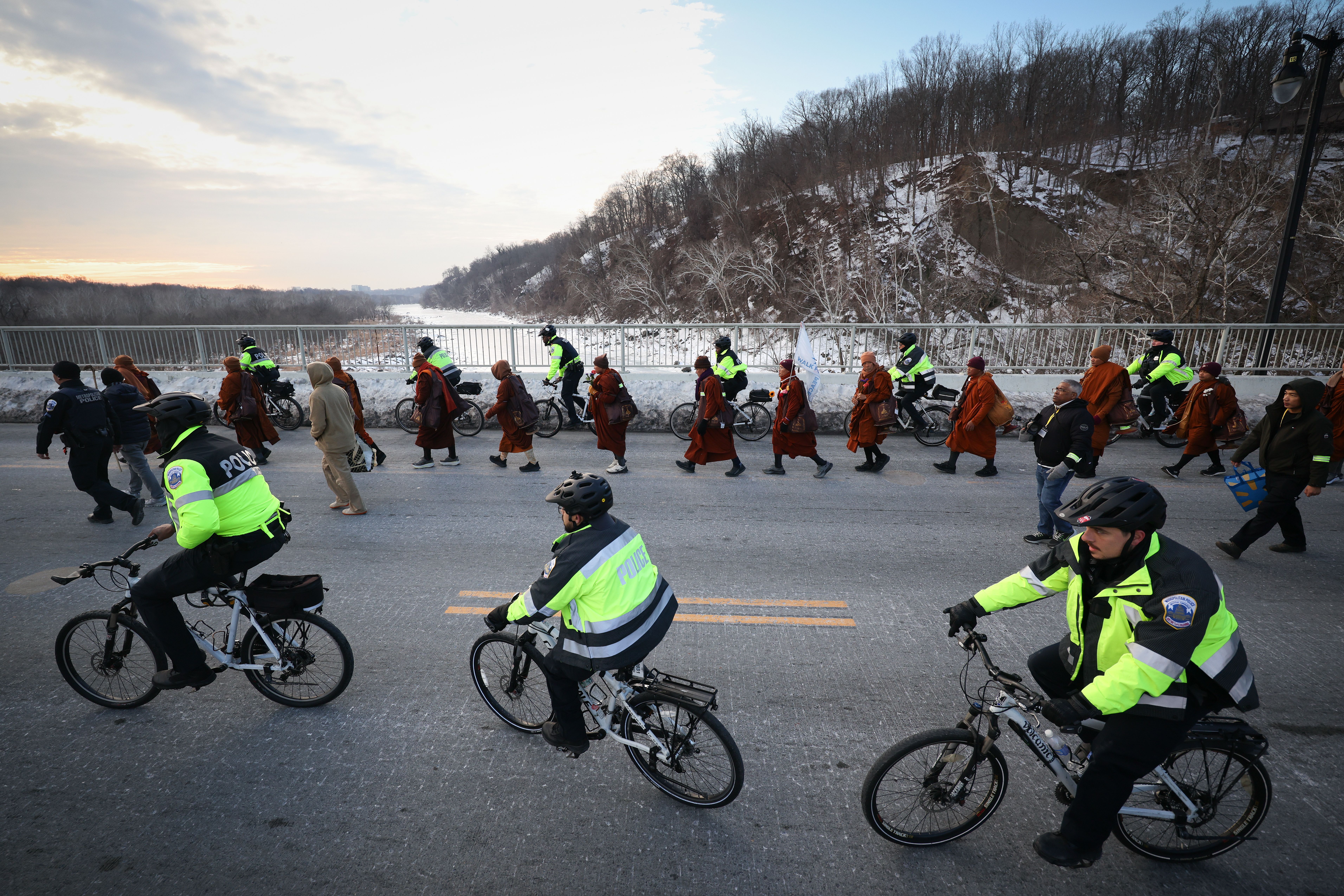 Police officers in bright yellow jackets on bicycles escort a line of monks in brown robes walking on a snowy road beside a river and wooded hill under a cloudy sky.
