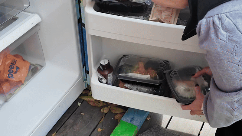 Person with black nail polish placing black plastic takeout containers into an open white refrigerator door on a wooden floor with scattered dry leaves.
