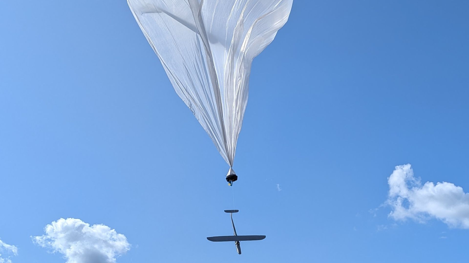 A white weather balloon in the sky carrying a small black glider beneath it with a clear blue sky and a few clouds in the background.