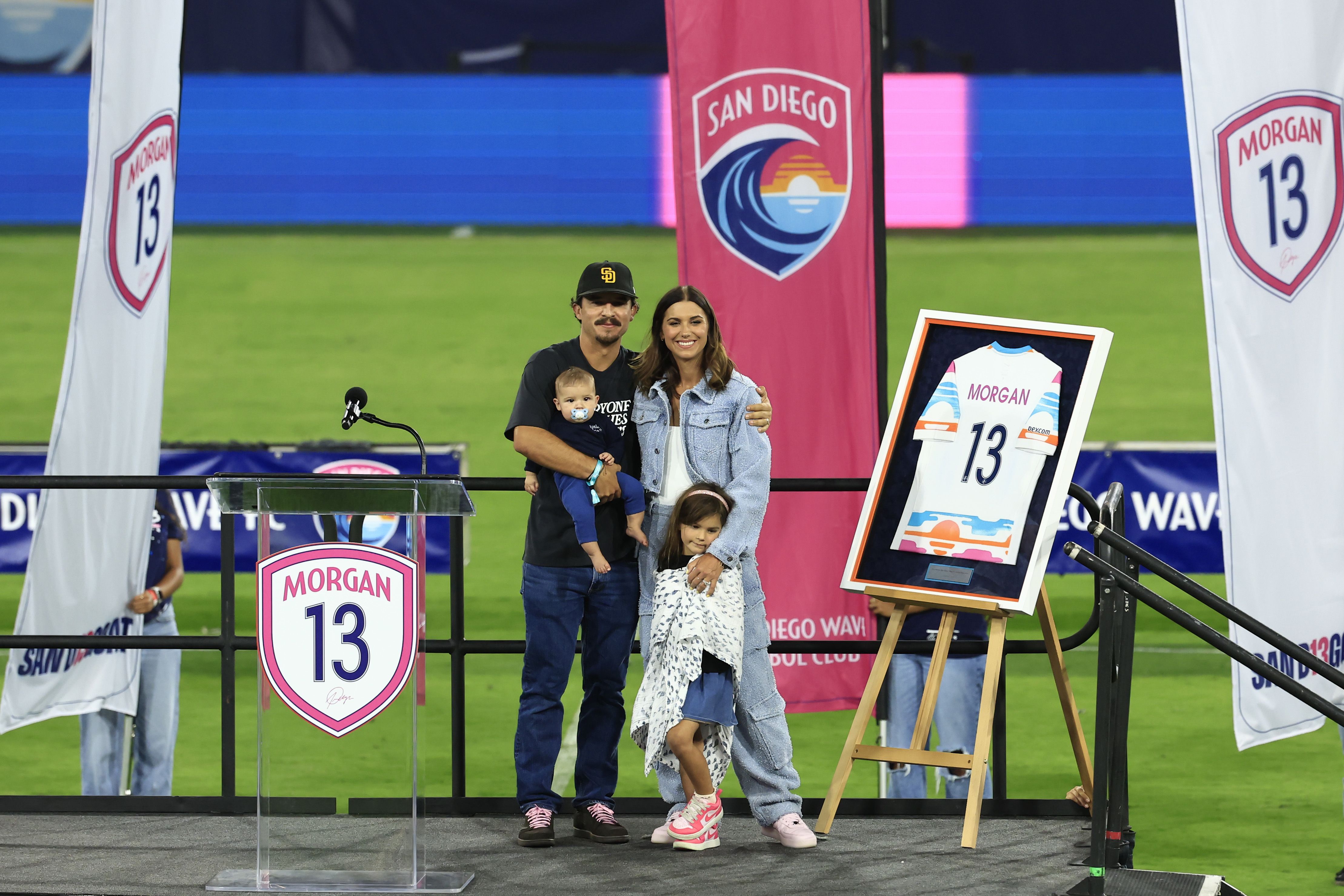 Alex Morgan stands with her family and a framed jersey on a stage during a ceremony on the field at Snapdragon Stadium.