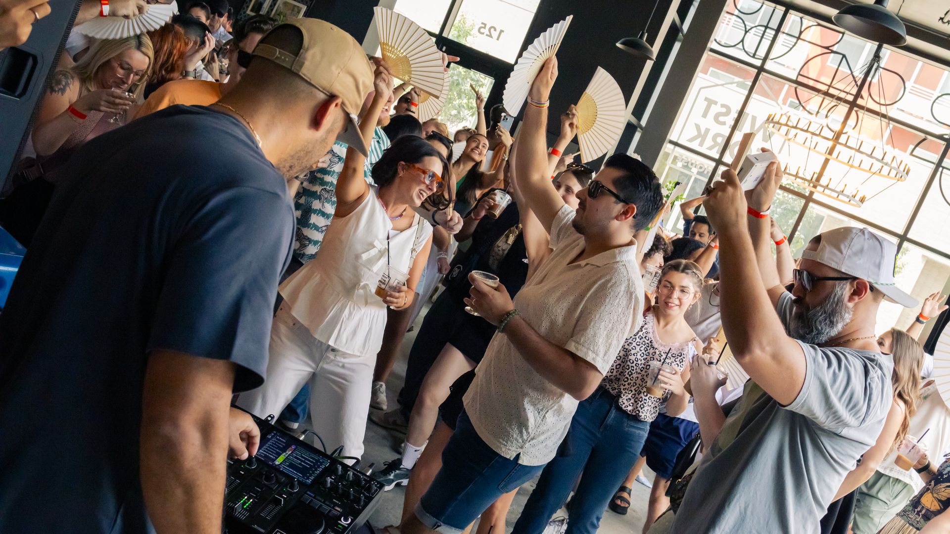 Crowd enjoying a lively indoor party with a DJ in a beige cap playing music. Several people hold drinks and white fans, dancing and smiling in a spacious, sunlit room.