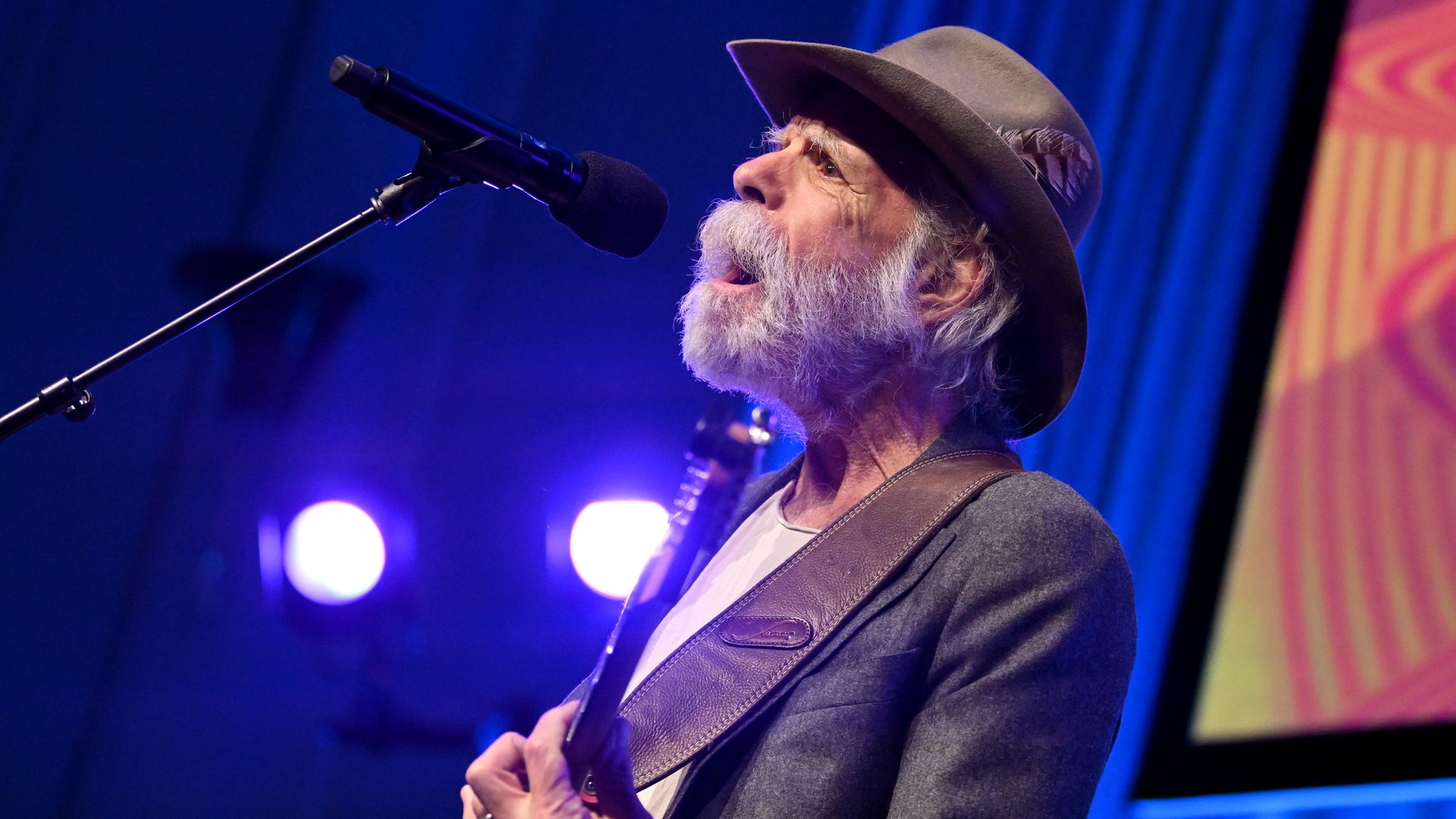 Elderly male musician with gray beard and hat singing into microphone and playing guitar on stage with blue and purple lighting.