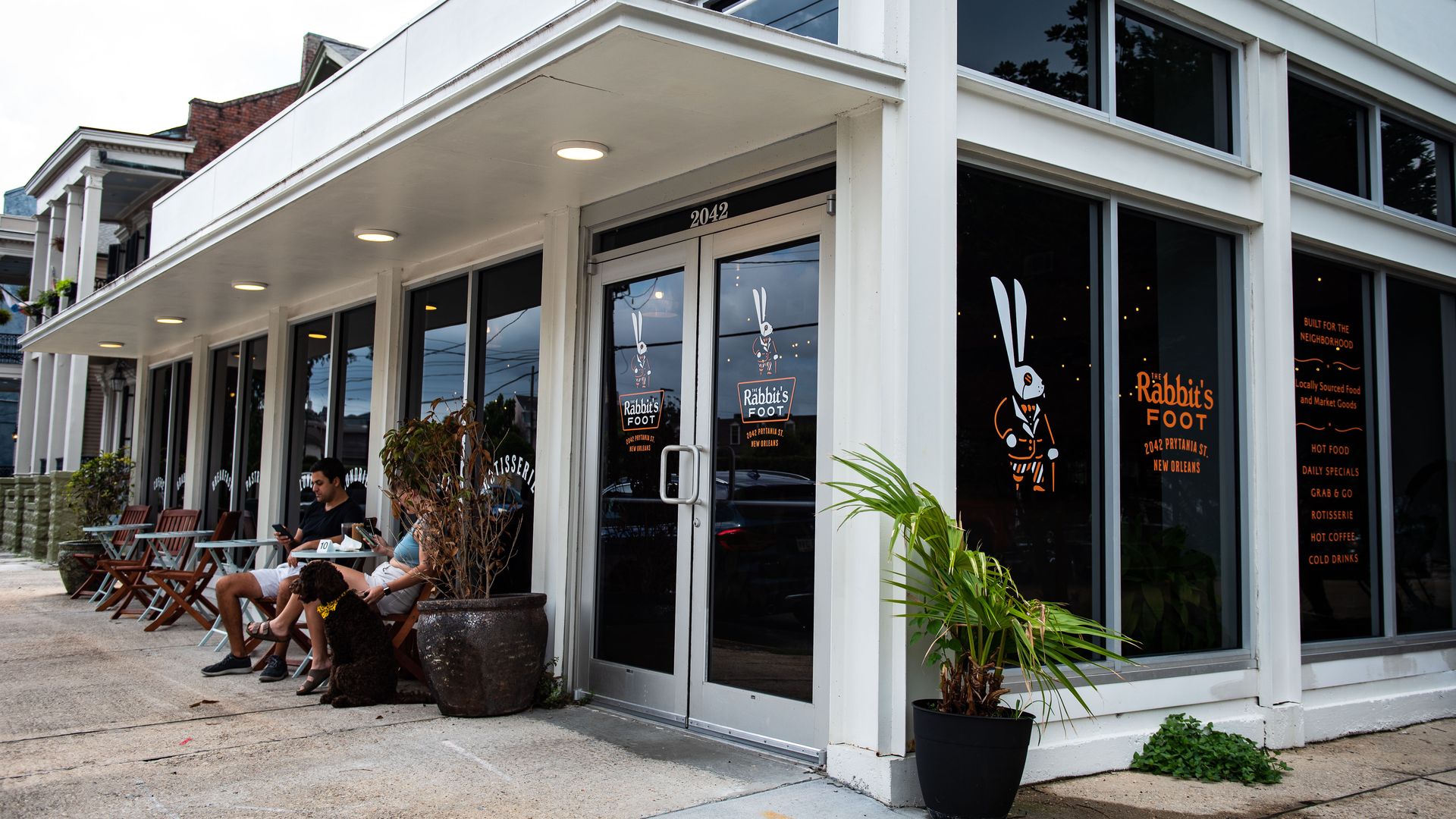 A view of Rabbit's Foot, a corner store bodega. The building is surrounded with glass windows and a couple patrons sit in chairs out front.
