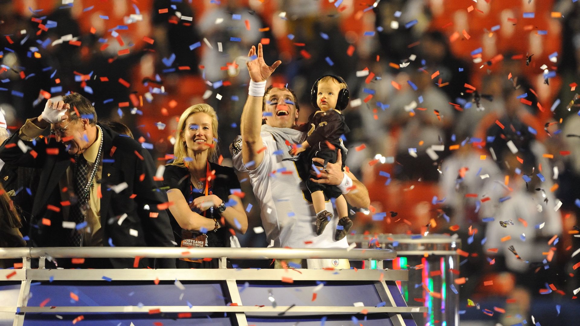 Drew Brees holds his son and stands next to his wife. Brees holds an arm into the air to catch confetti as it falls around them.