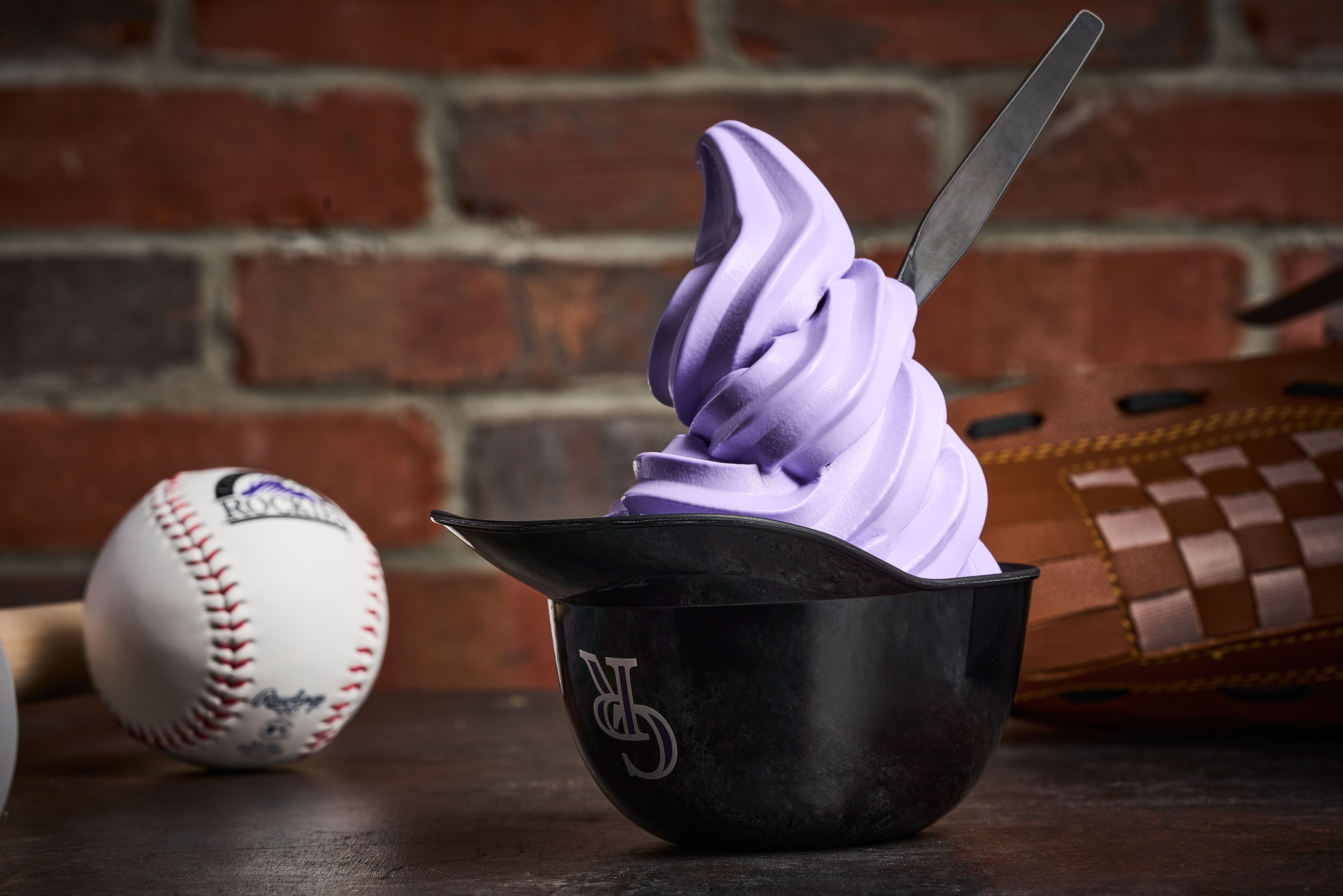 Purple soft-serve ice cream swirls up from a black top hat, with a long metal utensil stuck in it; a baseball and a brown glove lie on a wooden table before a brick wall.