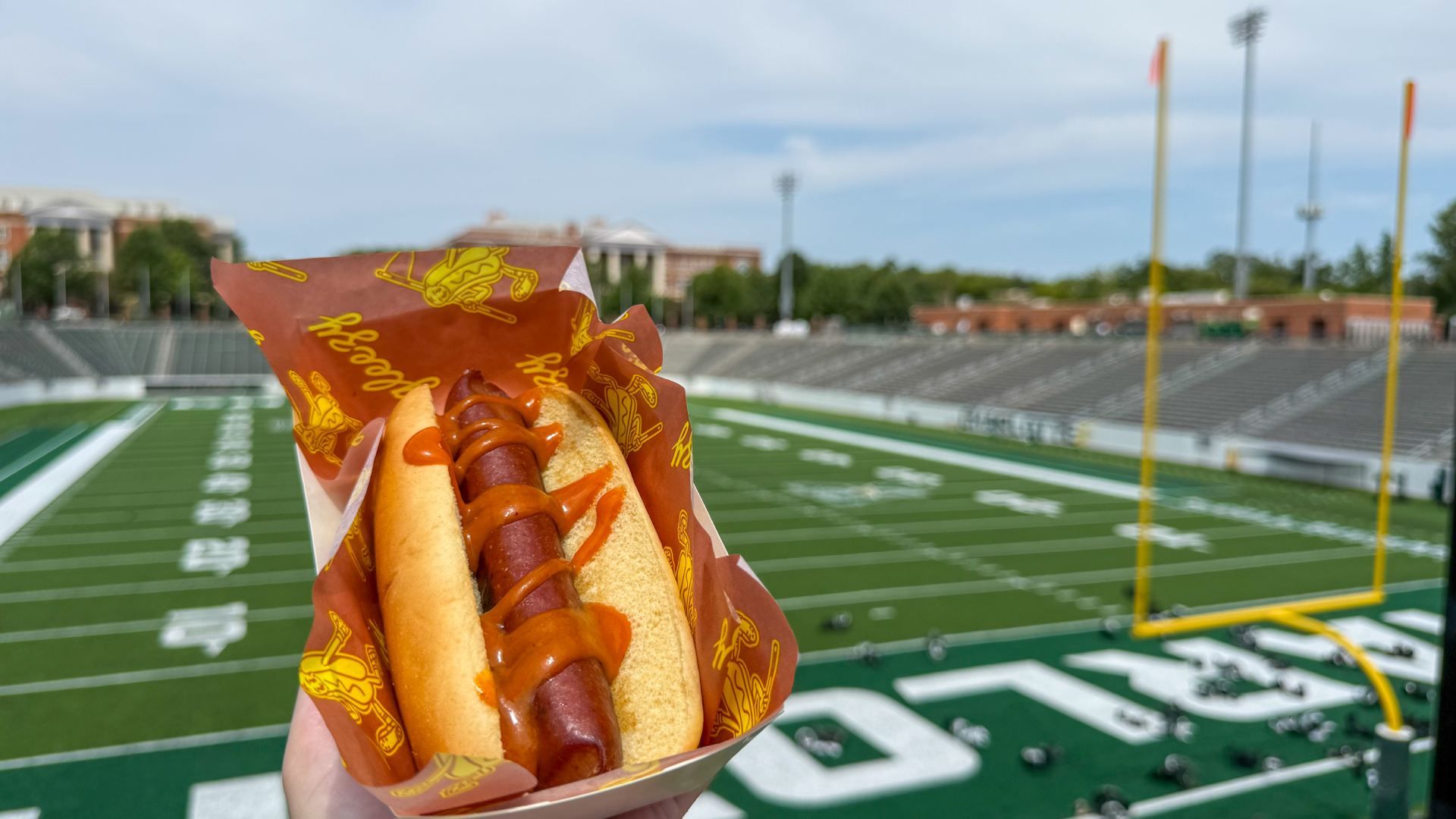 Hand holding a hot dog bun with ketchup and mustard, wrapped in orange paper with yellow logos, set against a green football field and stadium seating in the background.