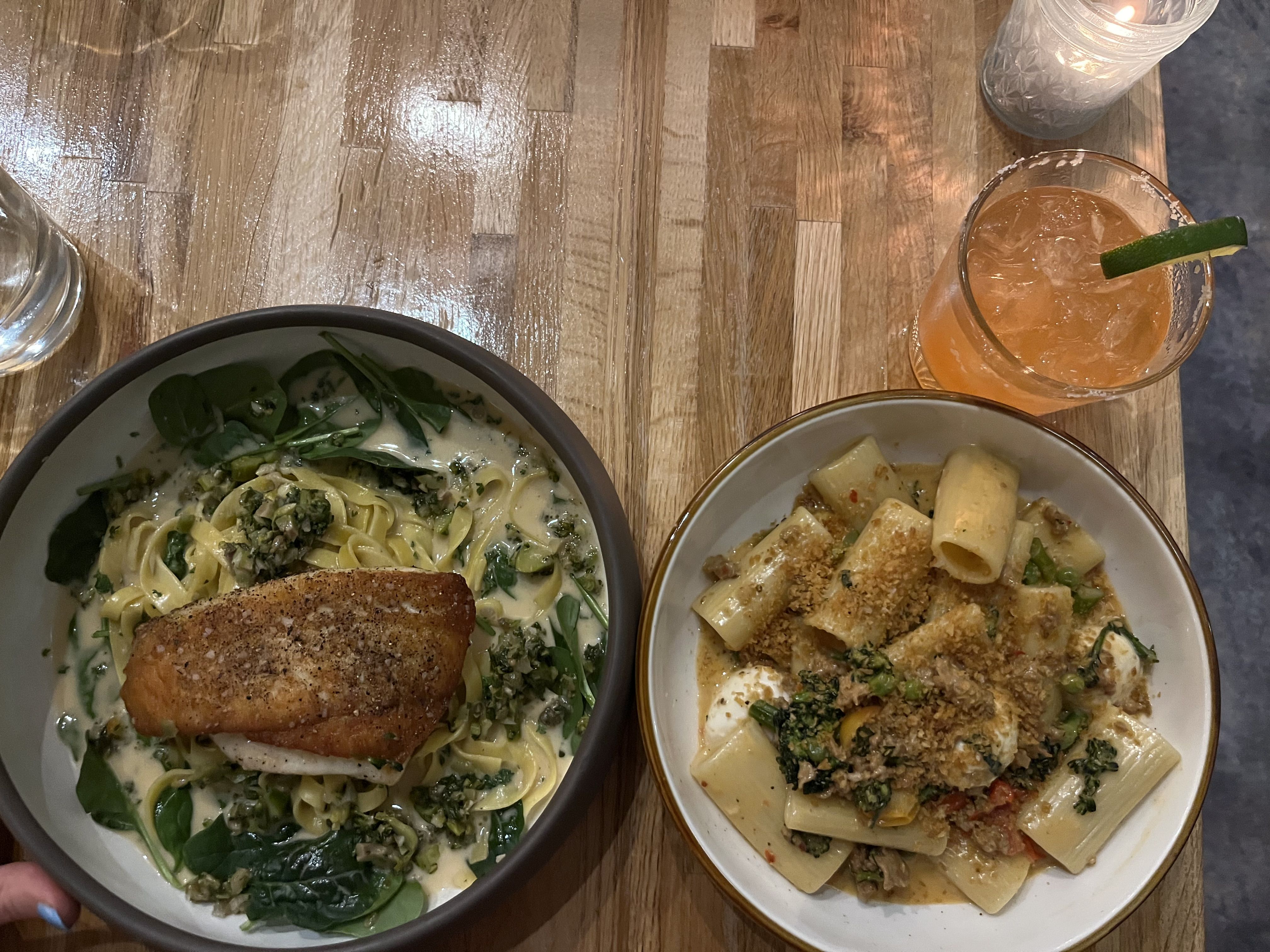 Top-down view of a wooden table with two pasta bowls: salmon on creamy spinach fettuccine, and rigatoni with meat sauce. An orange cocktail with lime and a lit candle nearby.