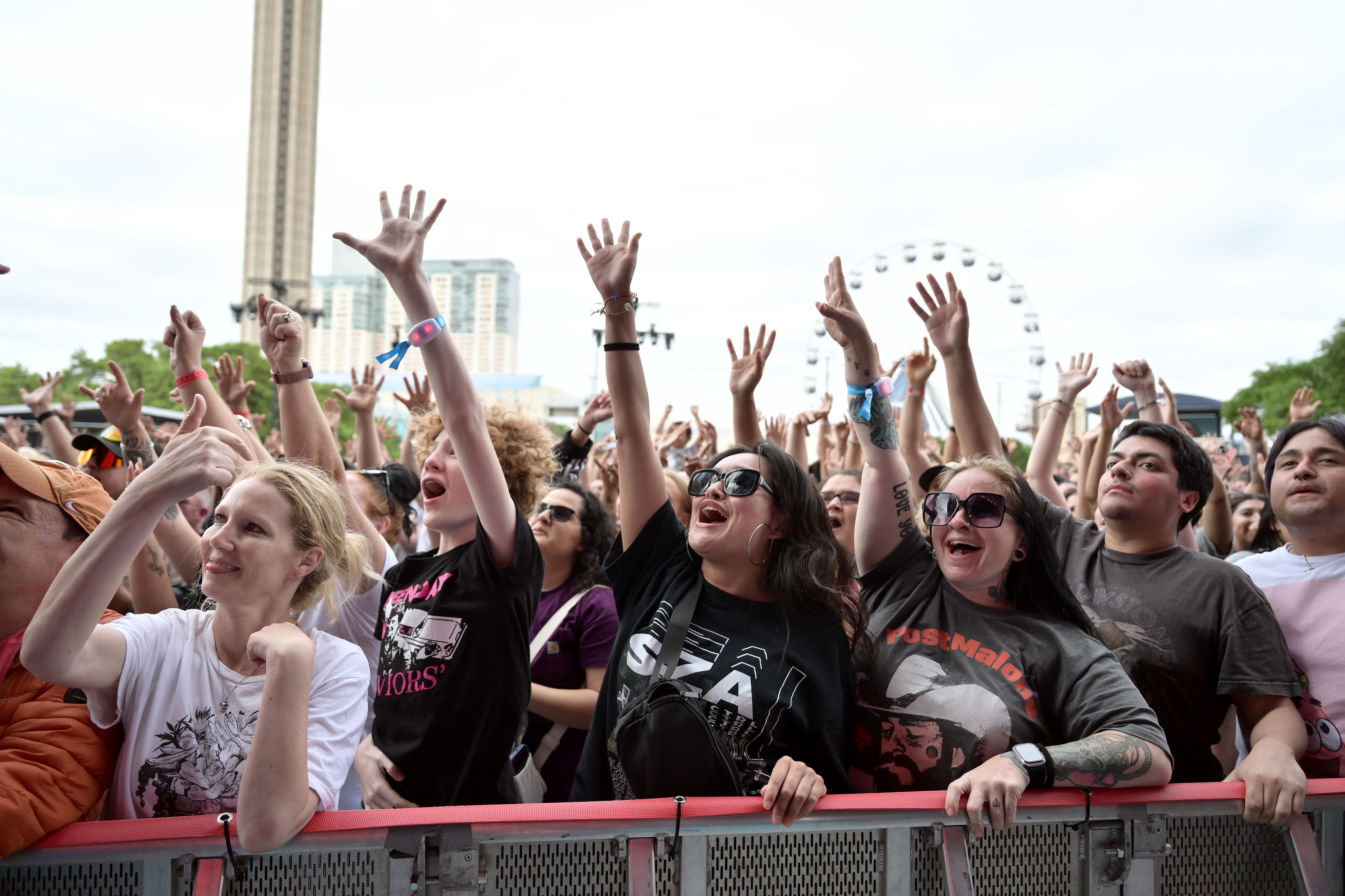 People enjoying the music festival with their hands in the air, with the Tower of the Americas and the Grand Hyatt in the background, as well as a ferris wheel.