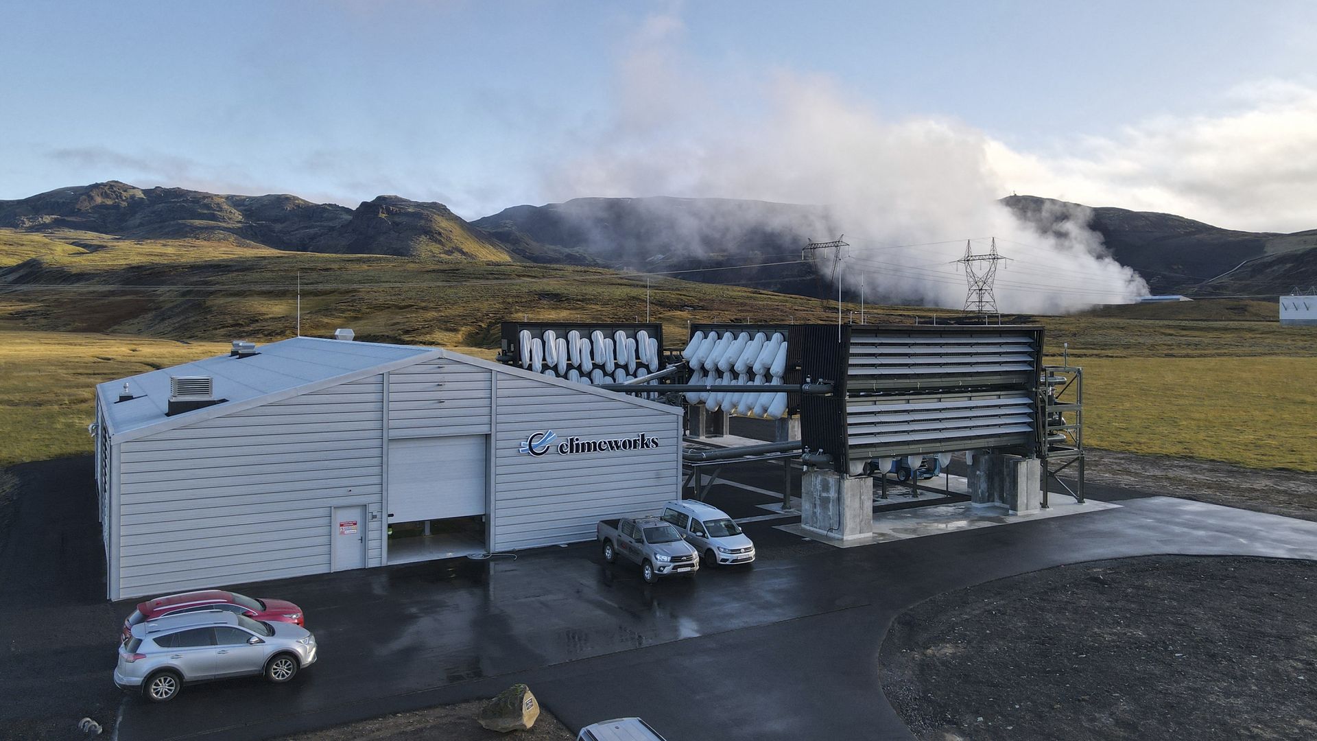 Photo of a factory with cars parked outside of it and steam rising in the distance.
