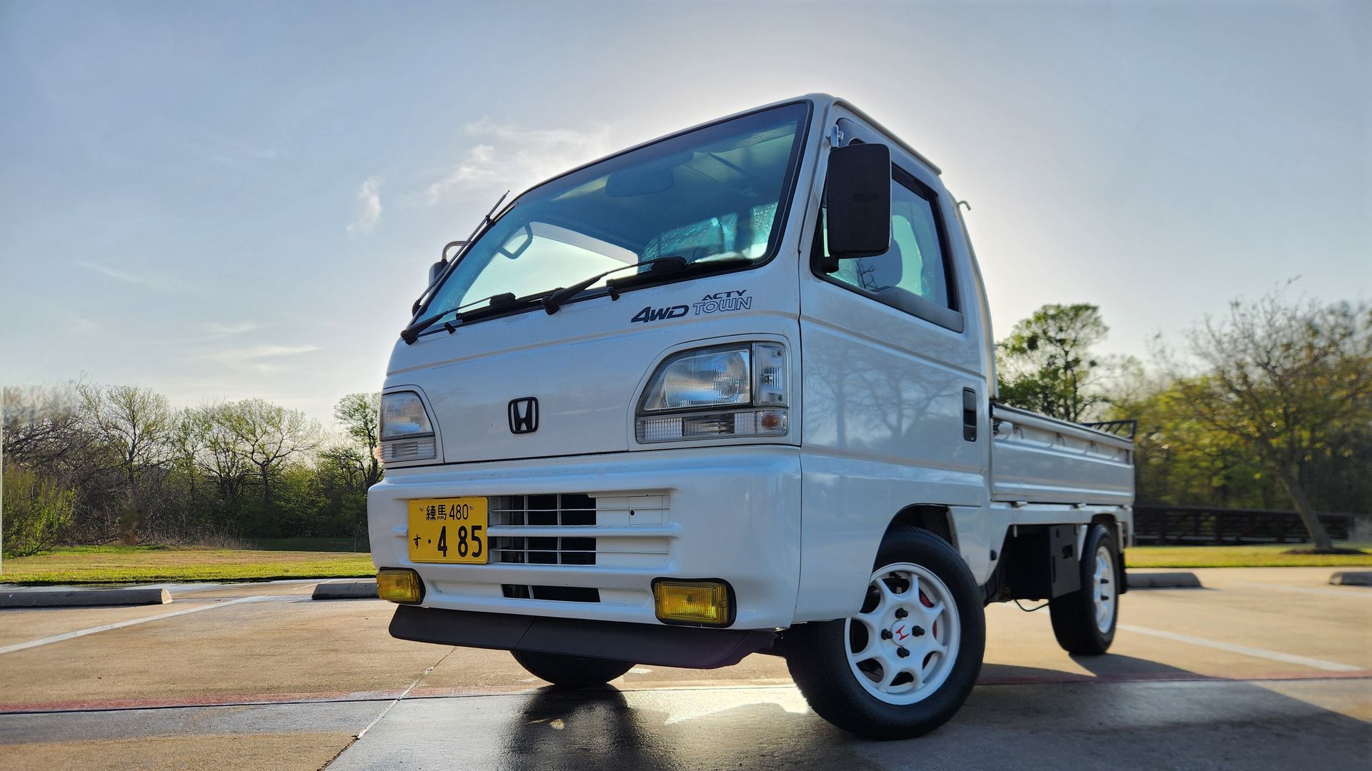 A small Japanese Honda truck, known as a kei truck, sits in a parking lot in front of trees. The small truck has an empty bed and is painted white. The sun sits behind behind the truck in a blue sky.