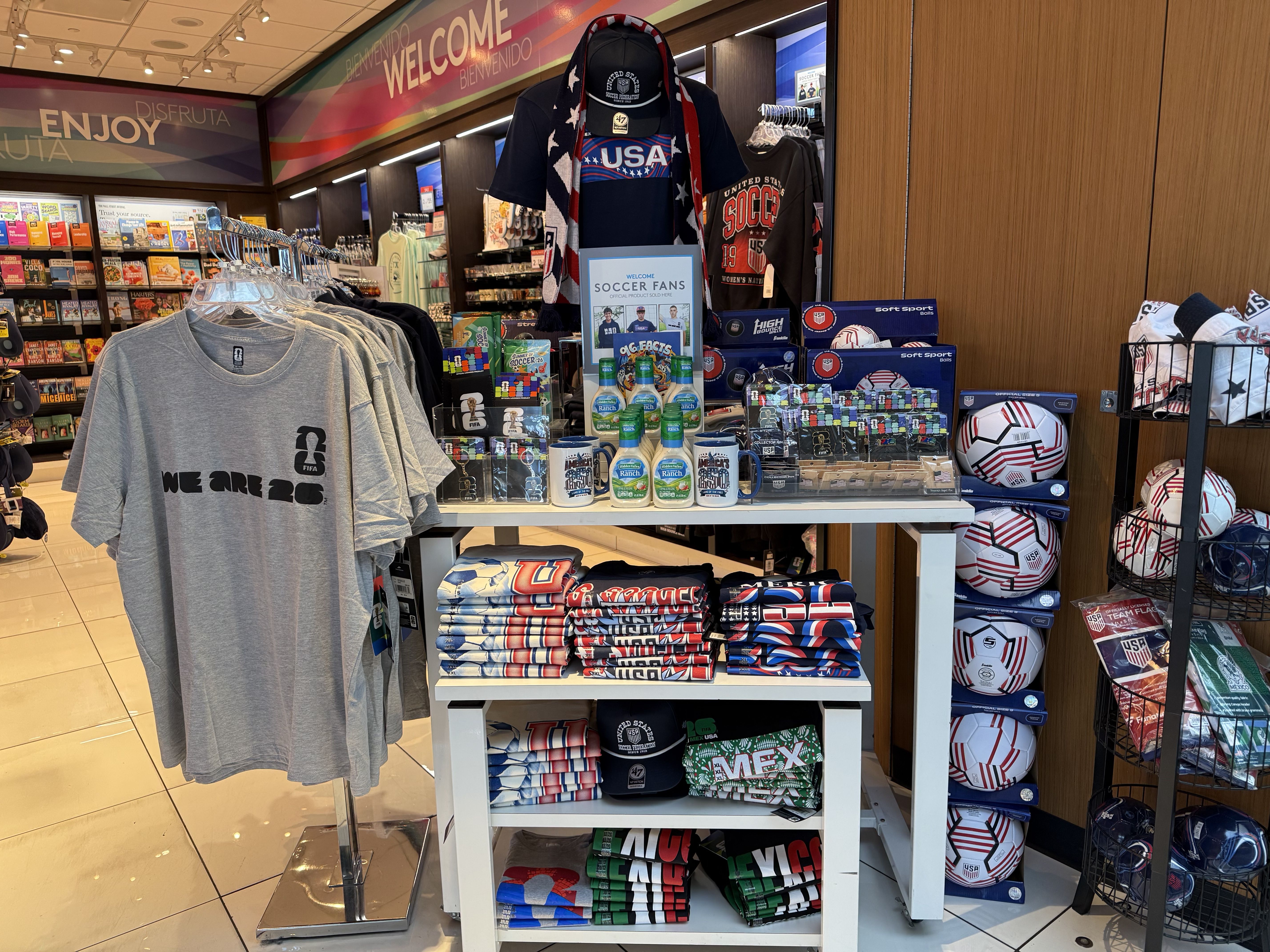 Souvenir shop scene with USA soccer gear: gray "WE ARE 26" tees in front, shelves of magazines to the left, a central "SOCCER FANS" display, and soccer balls to the right beneath a welcome banner.