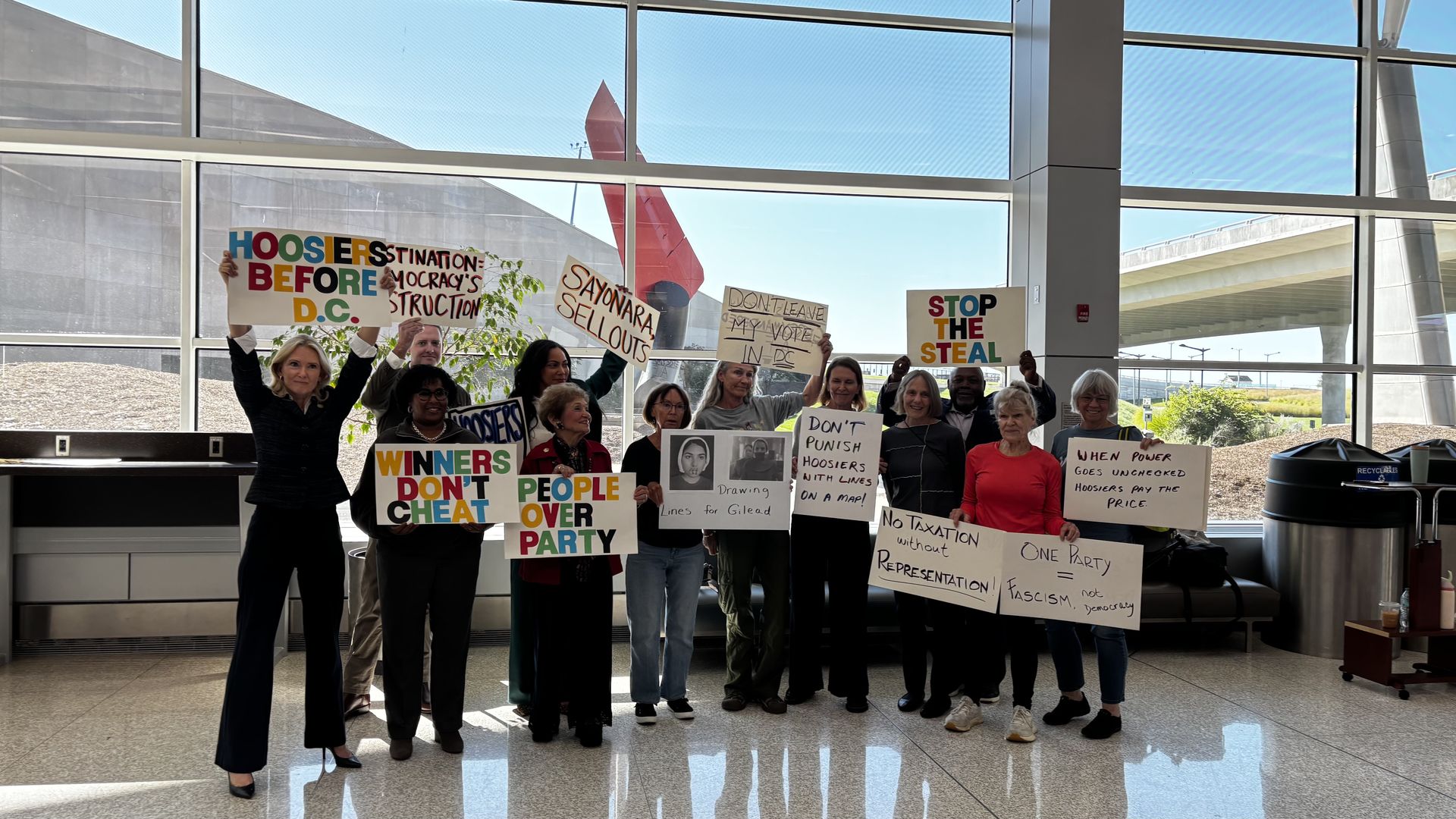 A group of diverse people indoors holding colorful protest signs with messages like "HOOSIERS BEFORE D.C.," "STOP THE STEAL," and "People Over Party" against political issues.
