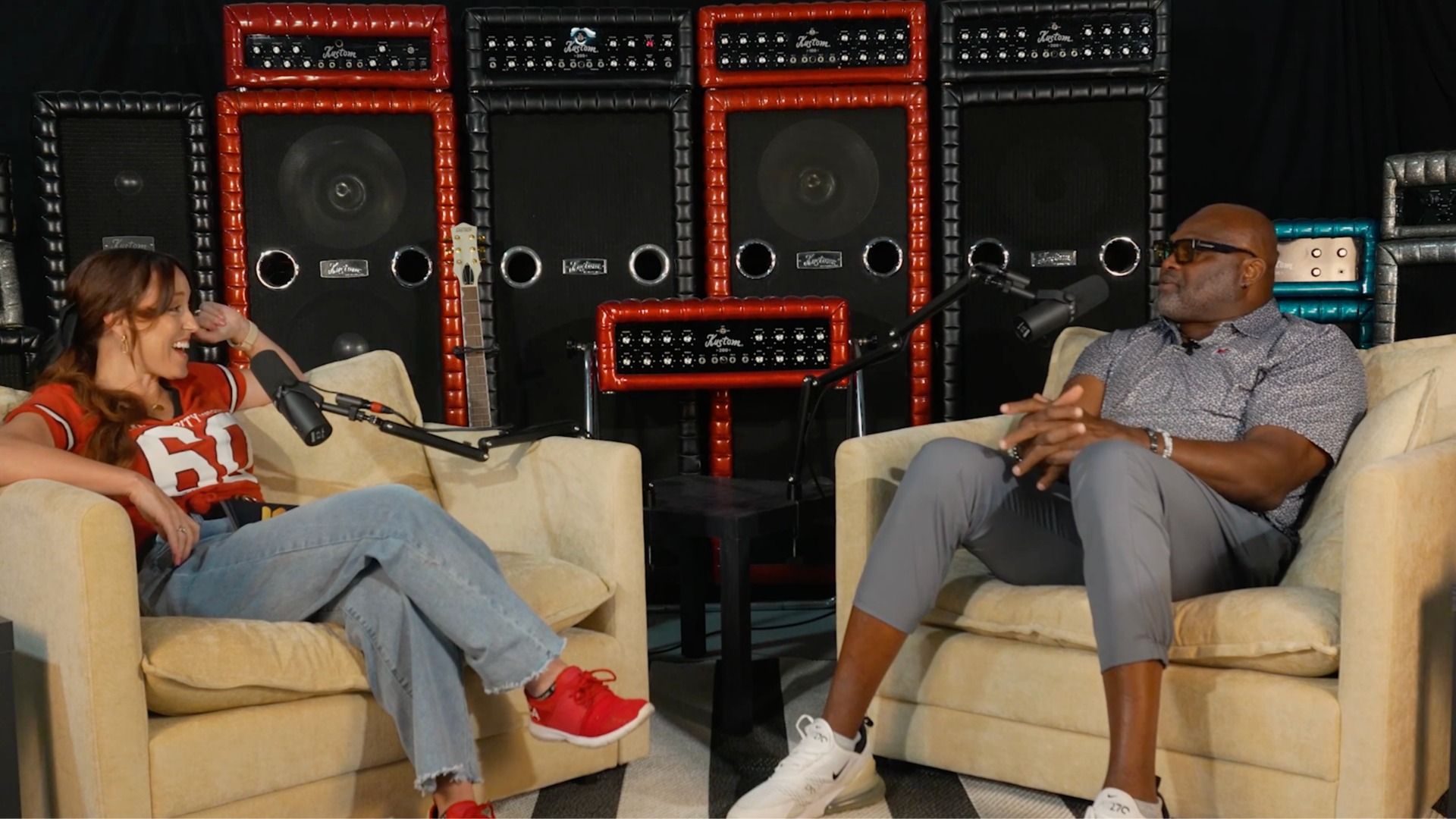 Two people sit on beige couches in a talk-show studio. The woman, Casi Joy, in a red jersey laughs into a microphone; the man, Neil Smith, in sunglasses listens. Behind them are red and black speaker stacks and a guitar.