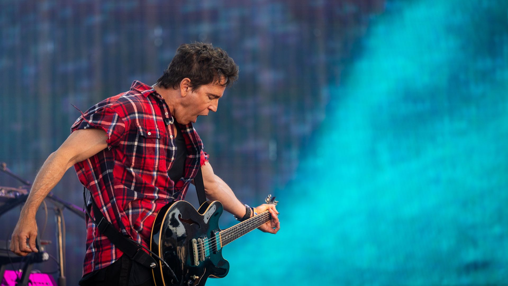 A guitarist in a red plaid shirt plays a black electric guitar on a stage with a blue-green LED backdrop.