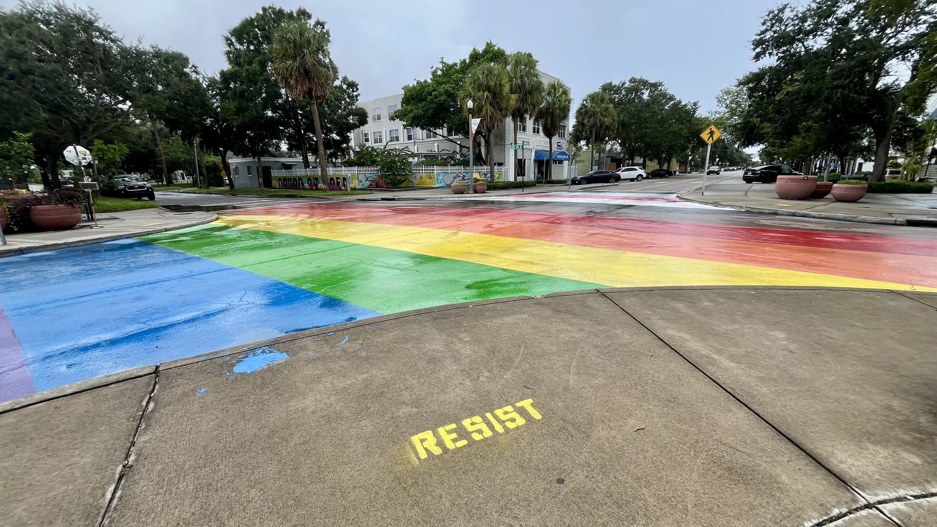 Colorful rainbow crosswalk on wet street with yellow stencil graffiti saying "RESIST" on sidewalk, surrounded by trees and buildings under gray sky.
