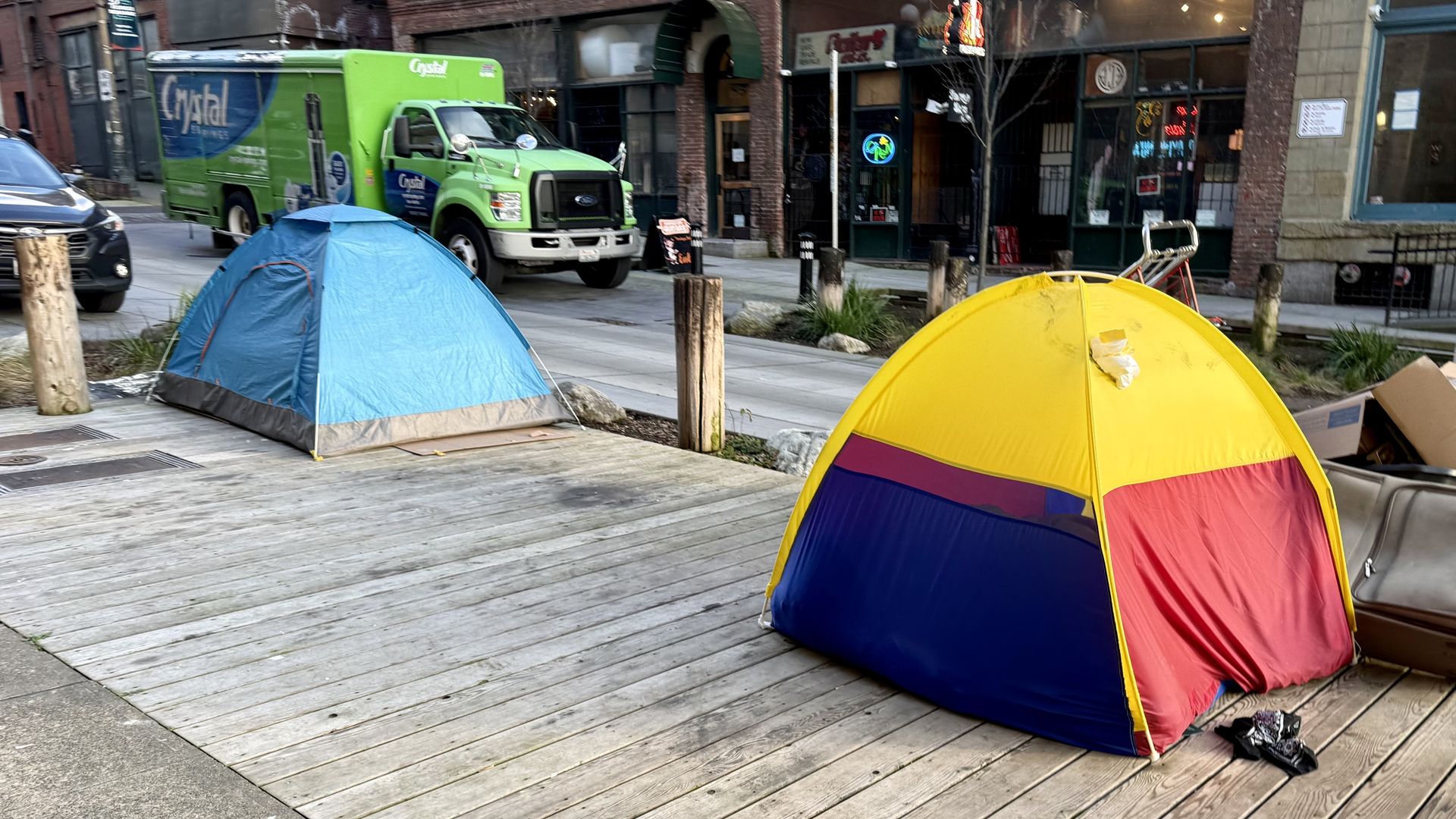 A city street scene with colorful camping tents on a wooden boardwalk: a blue tent on the left and a large yellow tent in the foreground, with a bright green delivery truck and brick buildings in the background.
