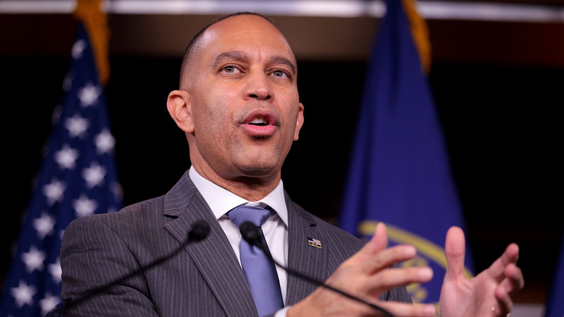 Hakeem Jeffries, wearing a pinstripe suit, standing in front of an American flag and another flag.