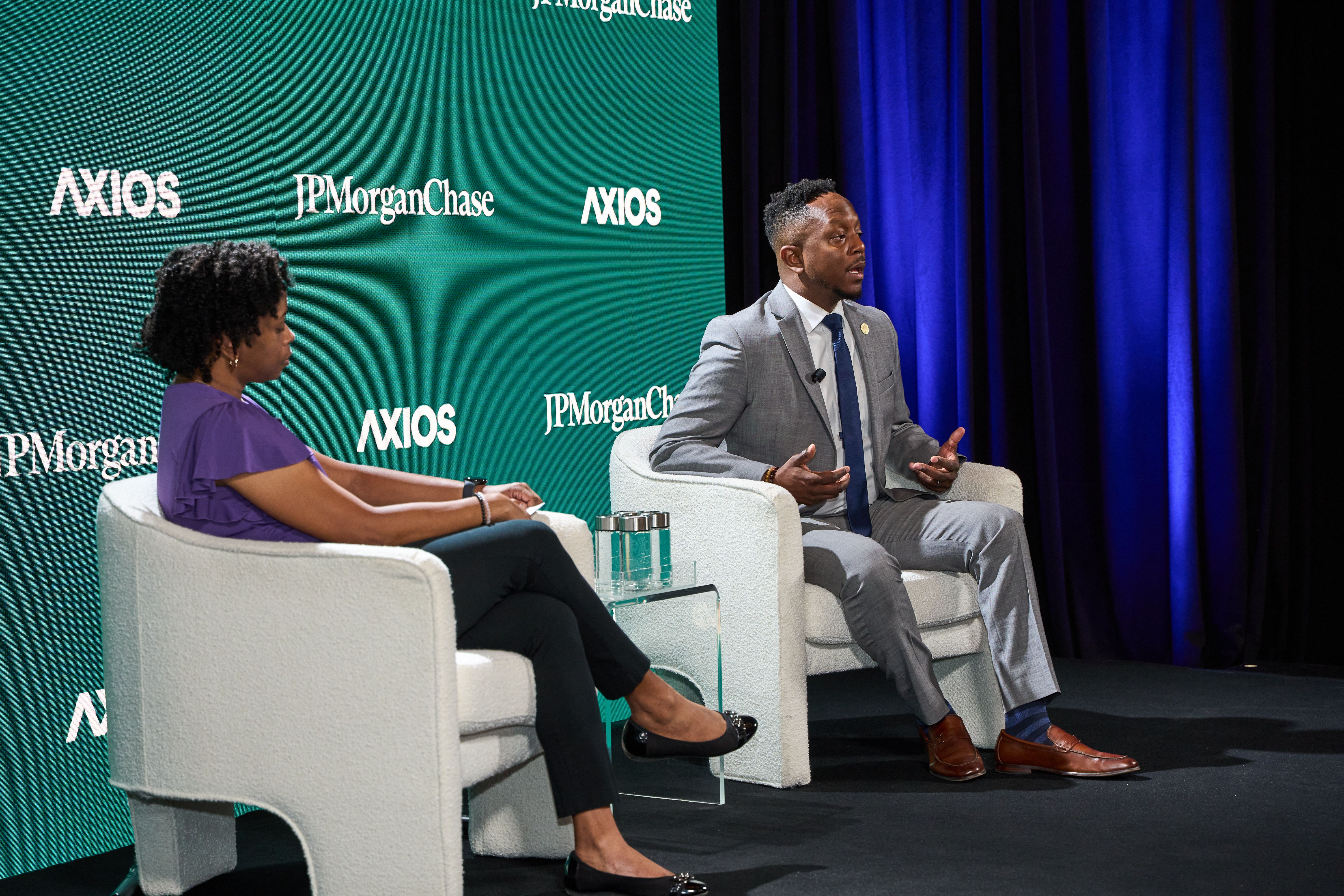 Two people seated in white chairs on stage during a discussion; man in gray suit with blue tie speaking, woman in purple blouse listening; backdrop shows Axios and JPMorgan Chase logos.