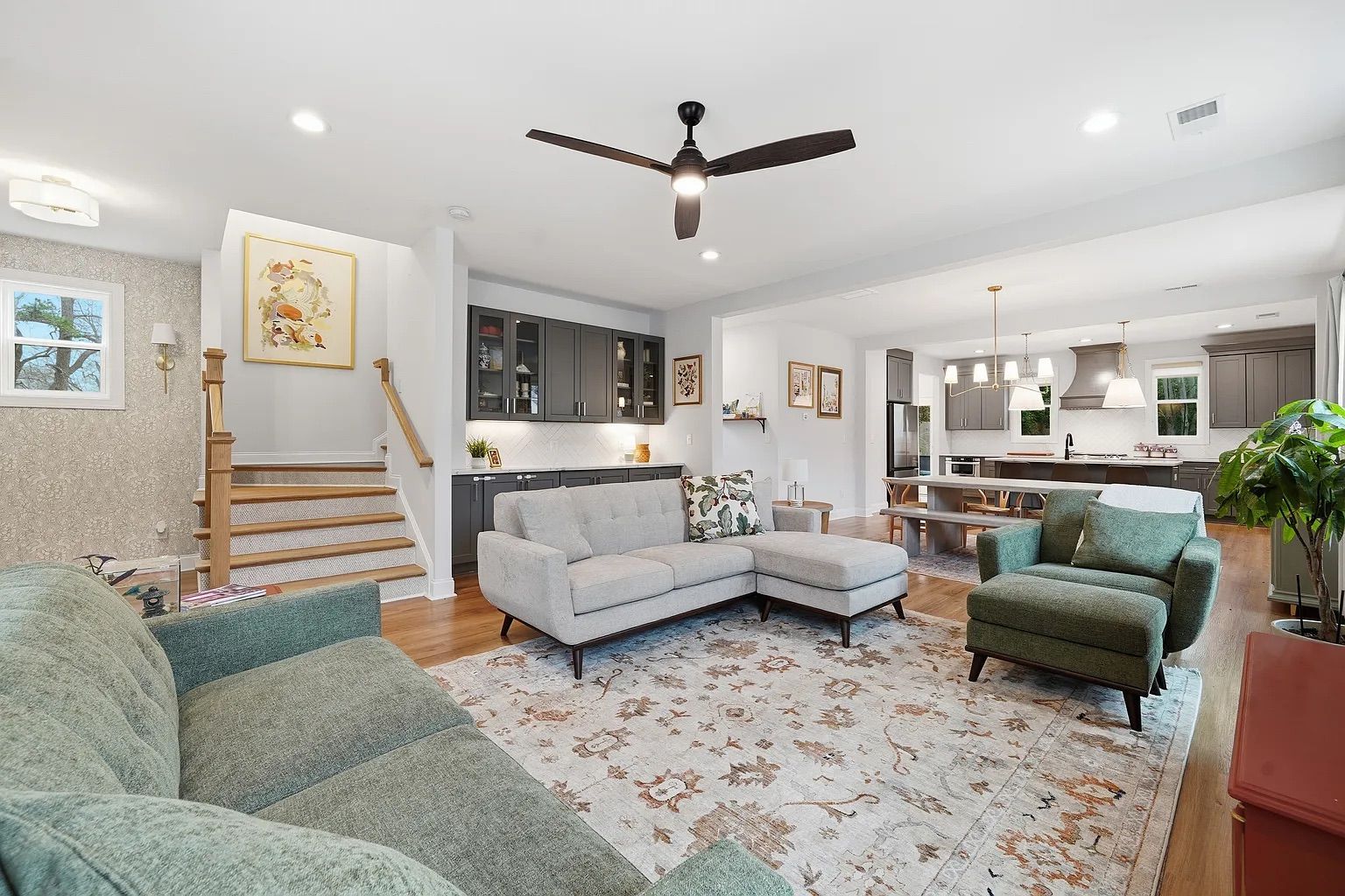 Spacious open living room with green and gray sofas on a patterned rug, wooden floors, stairs with wood railing, dark gray cabinets, and a modern kitchen with pendant lights in the background.
