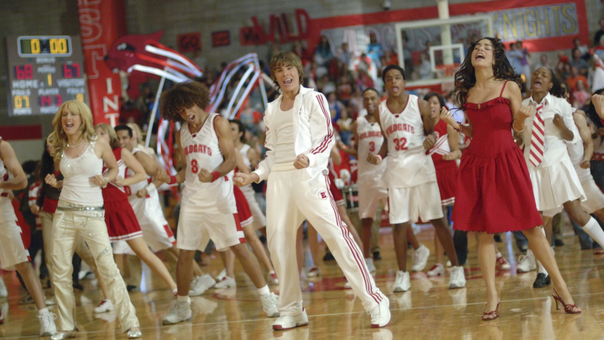 A group of high school students in Wildcats basketball uniforms, cheerleader outfits, and casual clothes are energetically celebrating on a gym floor with a scoreboard showing 68-67 in the background.