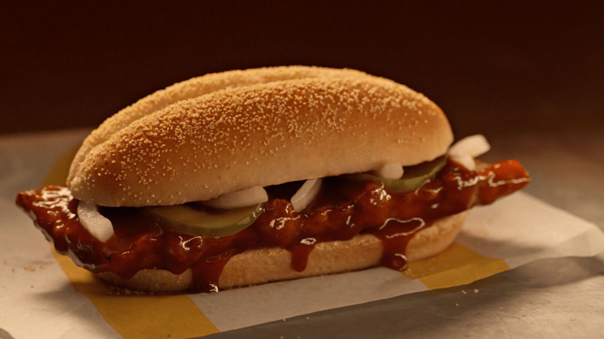 Barbecue sandwich with glazed meat, sliced pickles, and onions in a soft, golden-brown bun on wax paper.