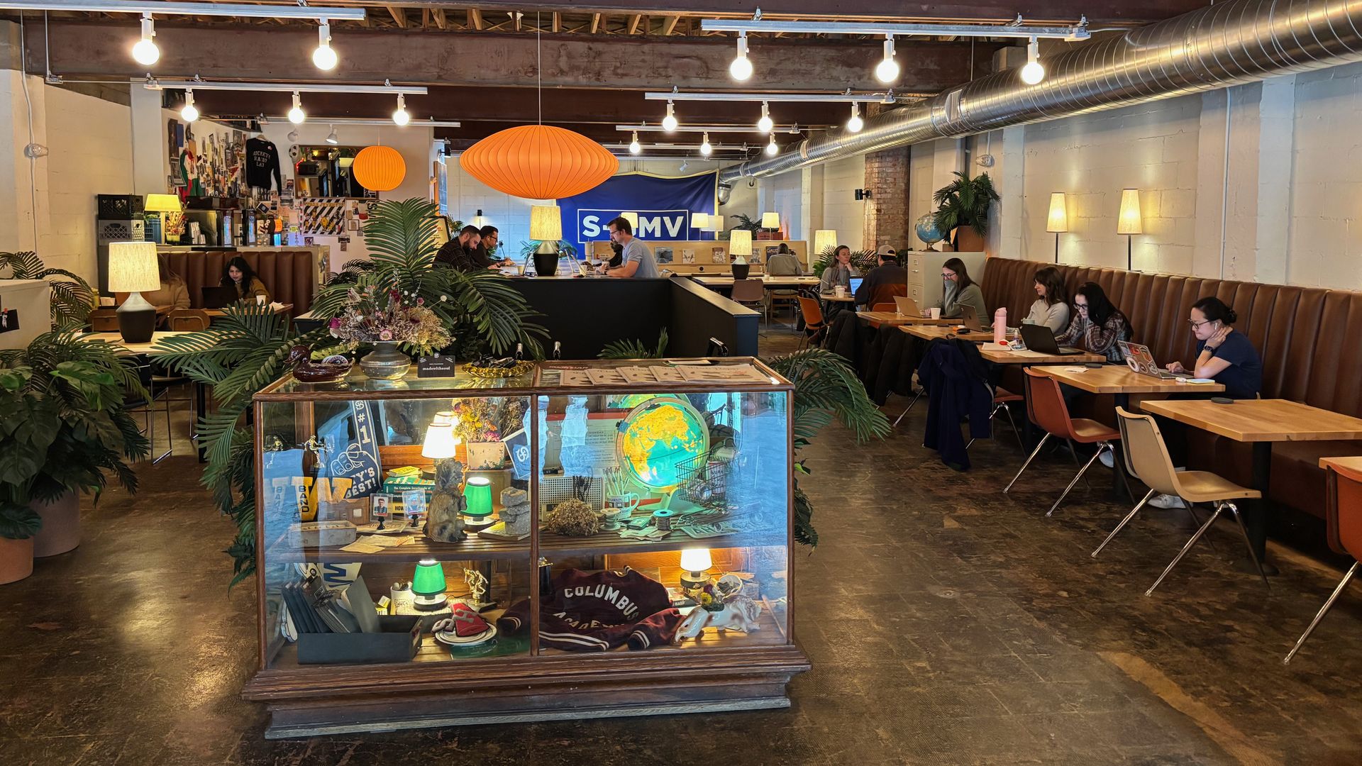 Industrial coworking cafe with exposed ceiling beams, string lights, and orange lanterns. A glass display cabinet sits in the foreground while people work at wooden tables along a long bench.